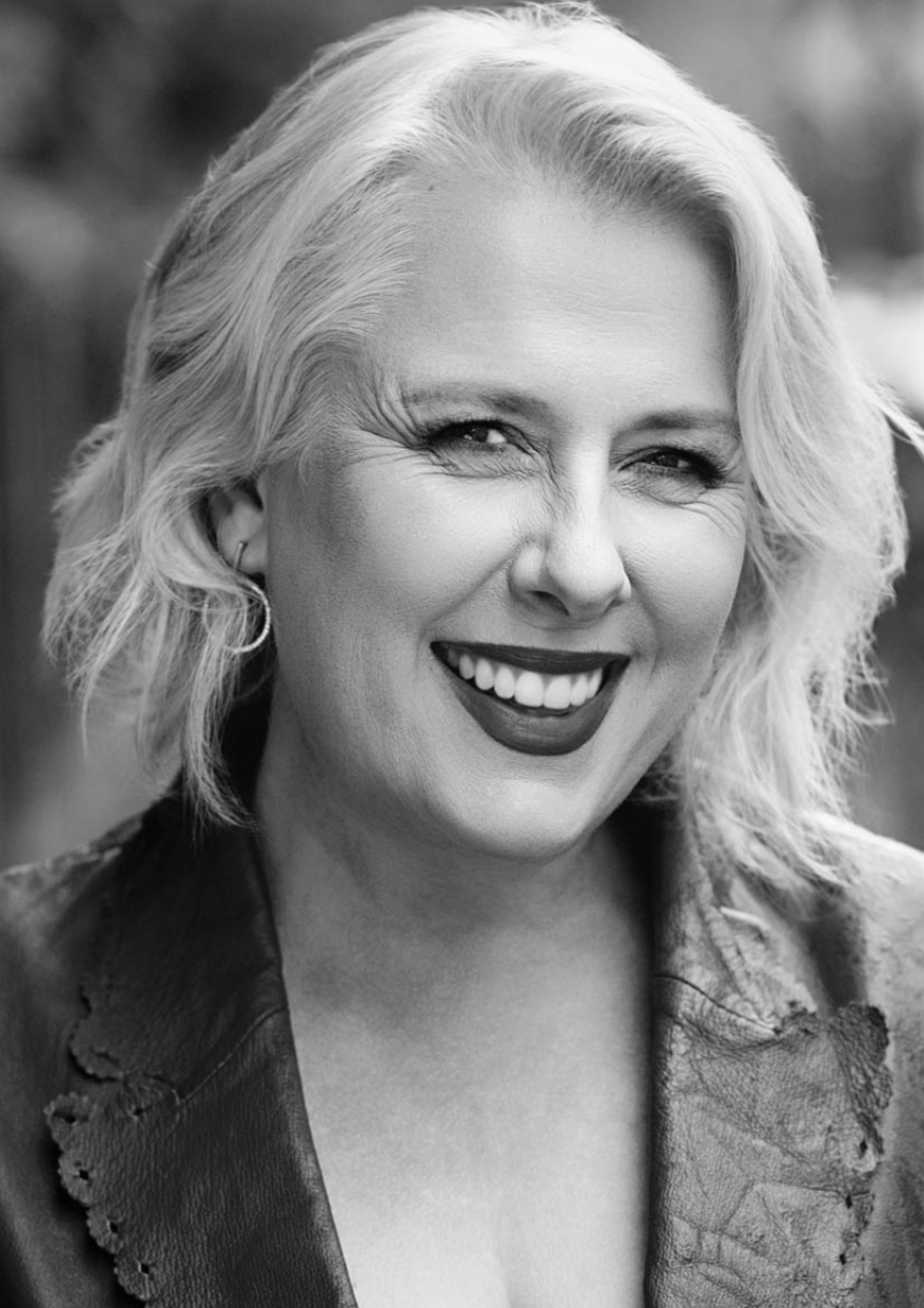A smiling woman with short, wavy blonde hair, wearing dark lipstick and a leather jacket, in a black-and-white portrait.