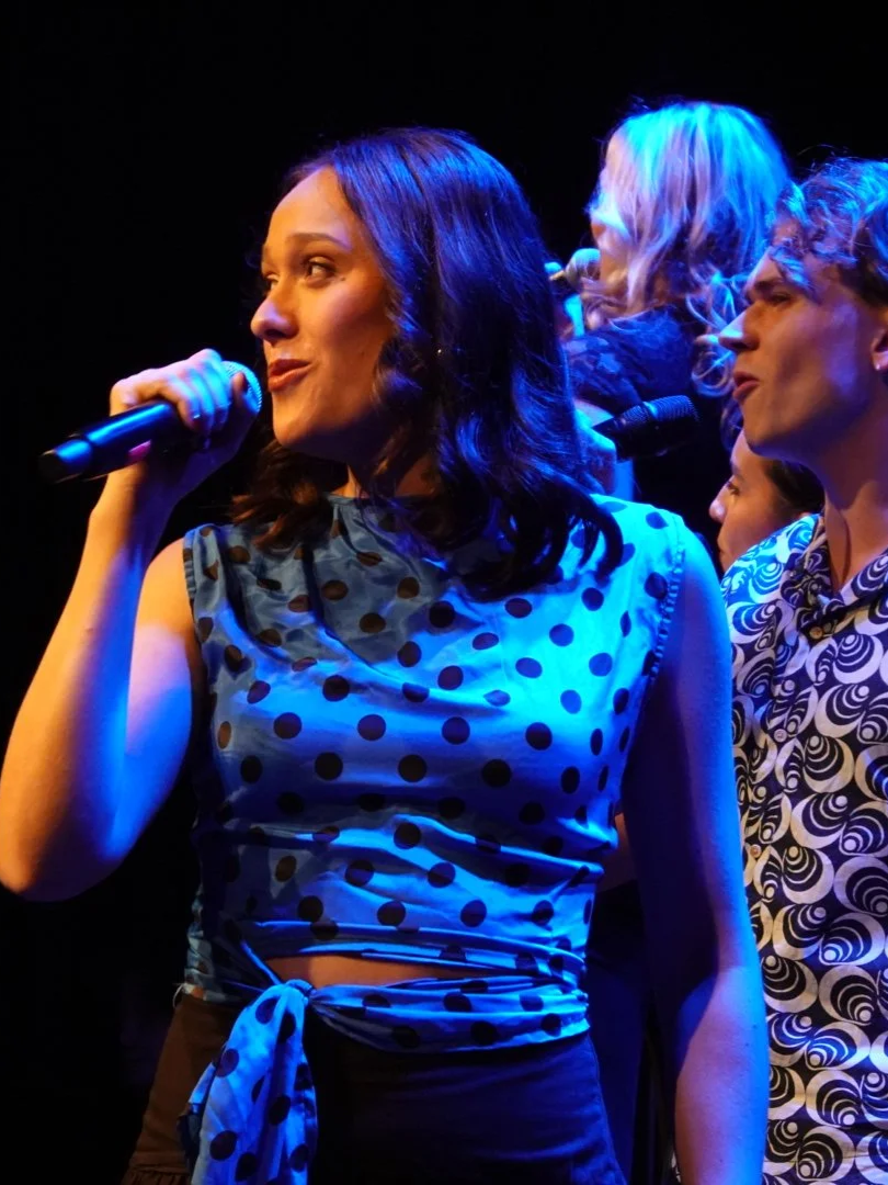 A woman with dark hair holding a microphone on stage, wearing a polka dot sleeveless top, with two women with blonde and curly hair and patterned shirts behind her.