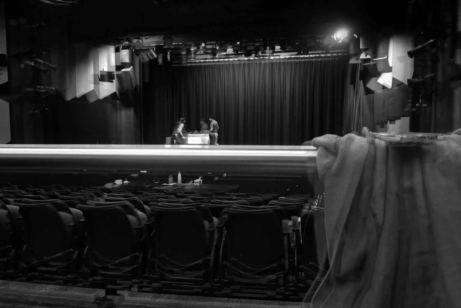 View of an empty theater or auditorium with a stage in the background, blackout curtains, and three people setting up equipment on stage; photo taken through a window, with a hand and some objects partially visible in the foreground.