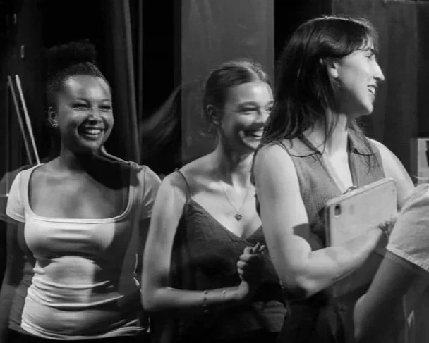 Three women standing in line, smiling and laughing, with a woman holding a smartphone.