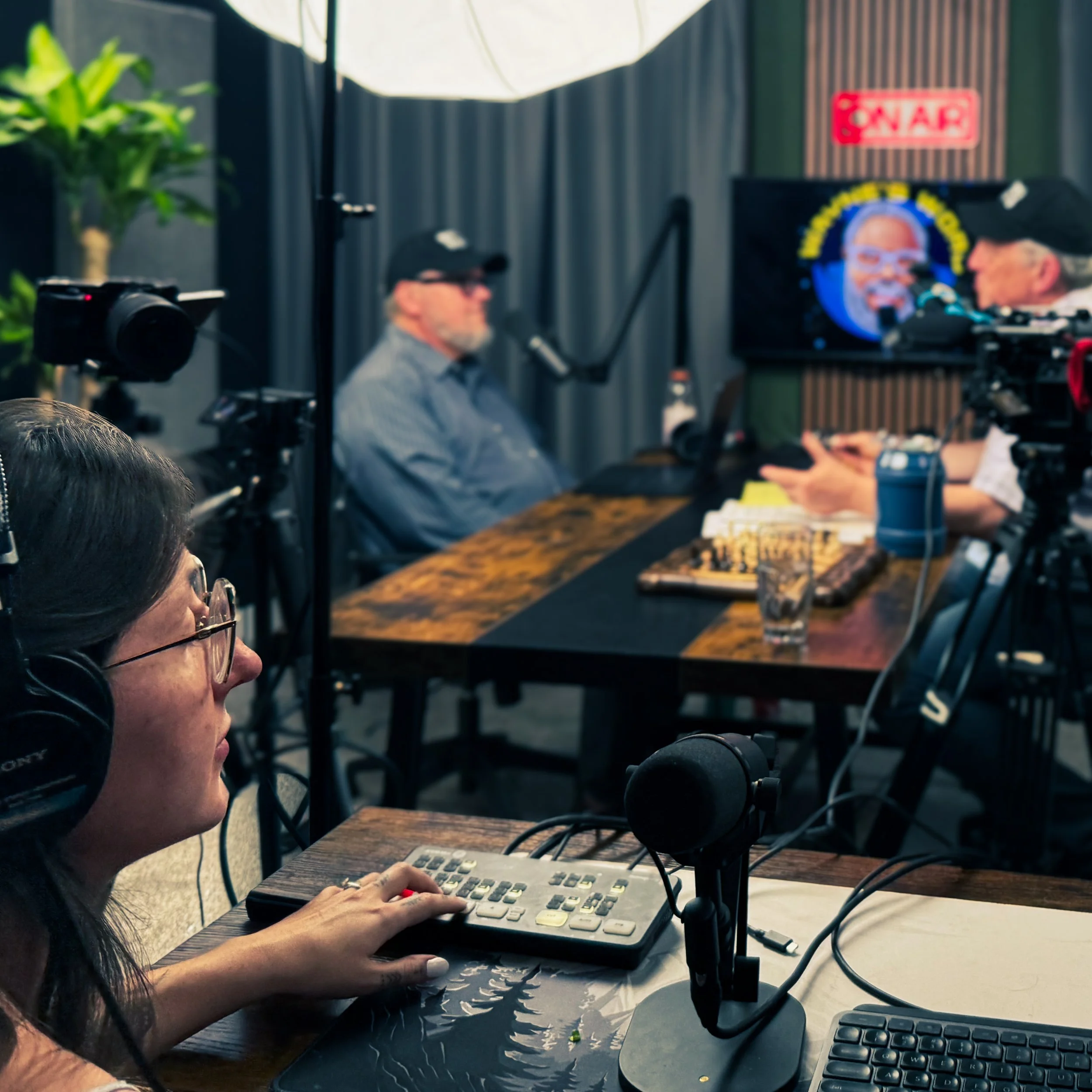 A woman with long dark hair and glasses working on a keyboard at a desk with a calculator, multiple computer monitors, and a microphone, in a studio setting.