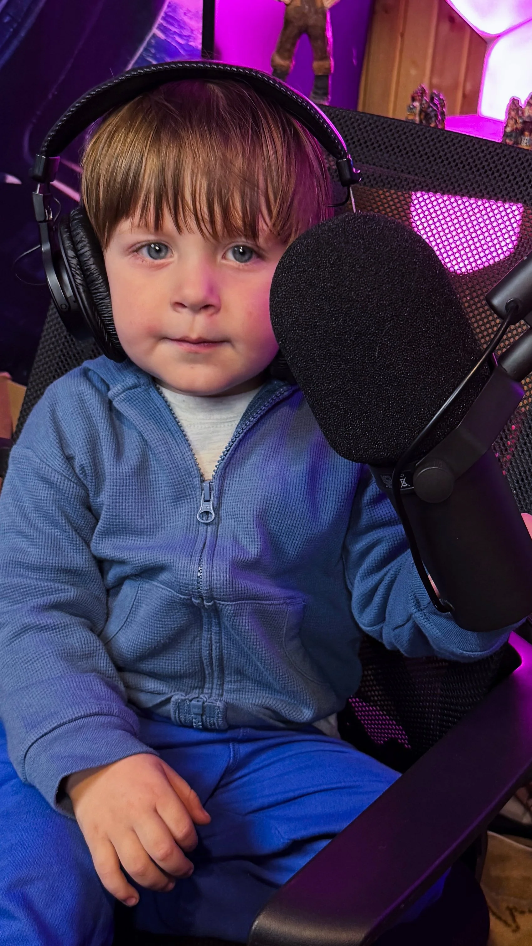 A young boy with brown hair and blue eyes, wearing a blue zip-up jacket, sitting in front of a microphone and headphones in a studio with colorful lighting.