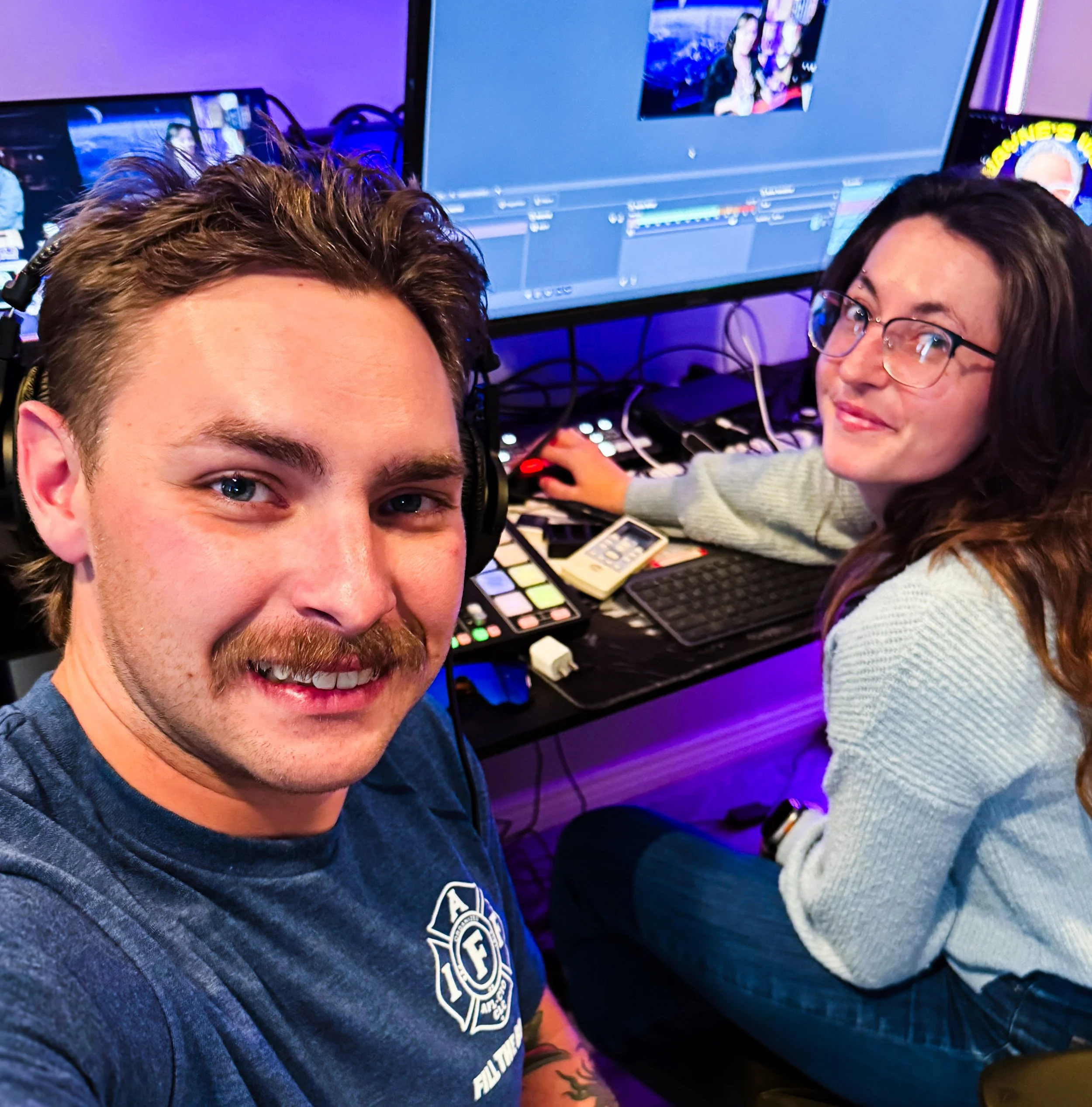Close-up of a young man's face with a beard and tattoos, wearing headphones and a dark shirt with a circular logo, in a room with purple lighting and computer equipment, including monitors and electronic music production gear, with a young woman working on a computer in the background.