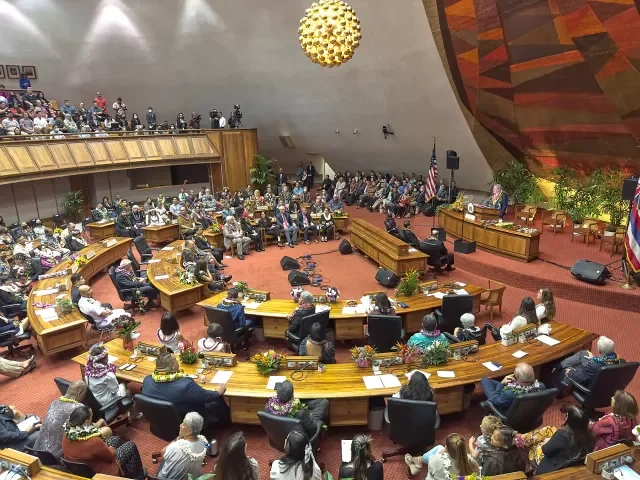 A large assembly hall filled with seated people, many wearing leis, arranged around curved desks, with a stage, American flags, and large decorative ceiling element.