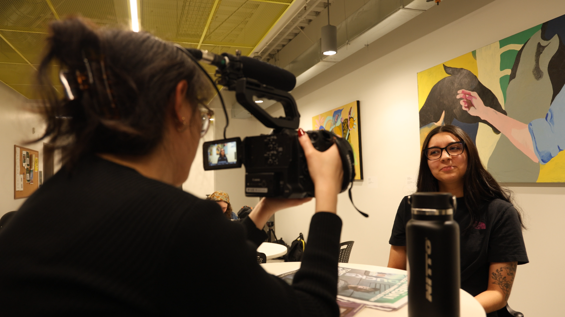 A woman with dark hair and glasses being filmed with a professional video camera at an art gallery or museum. The background features colorful artwork on the wall.