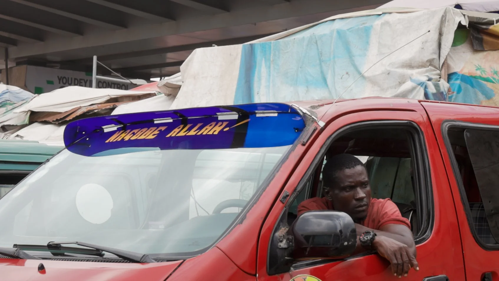A man with a serious expression sitting inside a red emergency vehicle labeled 'Mobile AllAfrica', parked in an area with makeshift shelters and signs in the background.