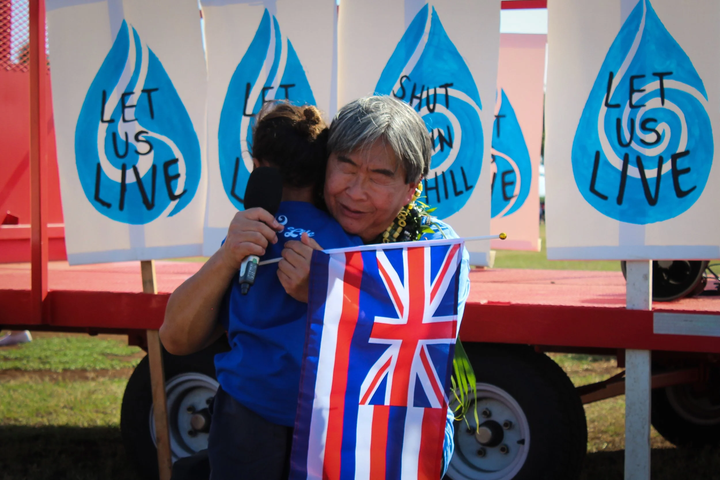Two people hugging at an outdoor event, one person is holding an Australian flag. Behind them are blue banners with a water droplet shape and the words 'Let us shut down'.