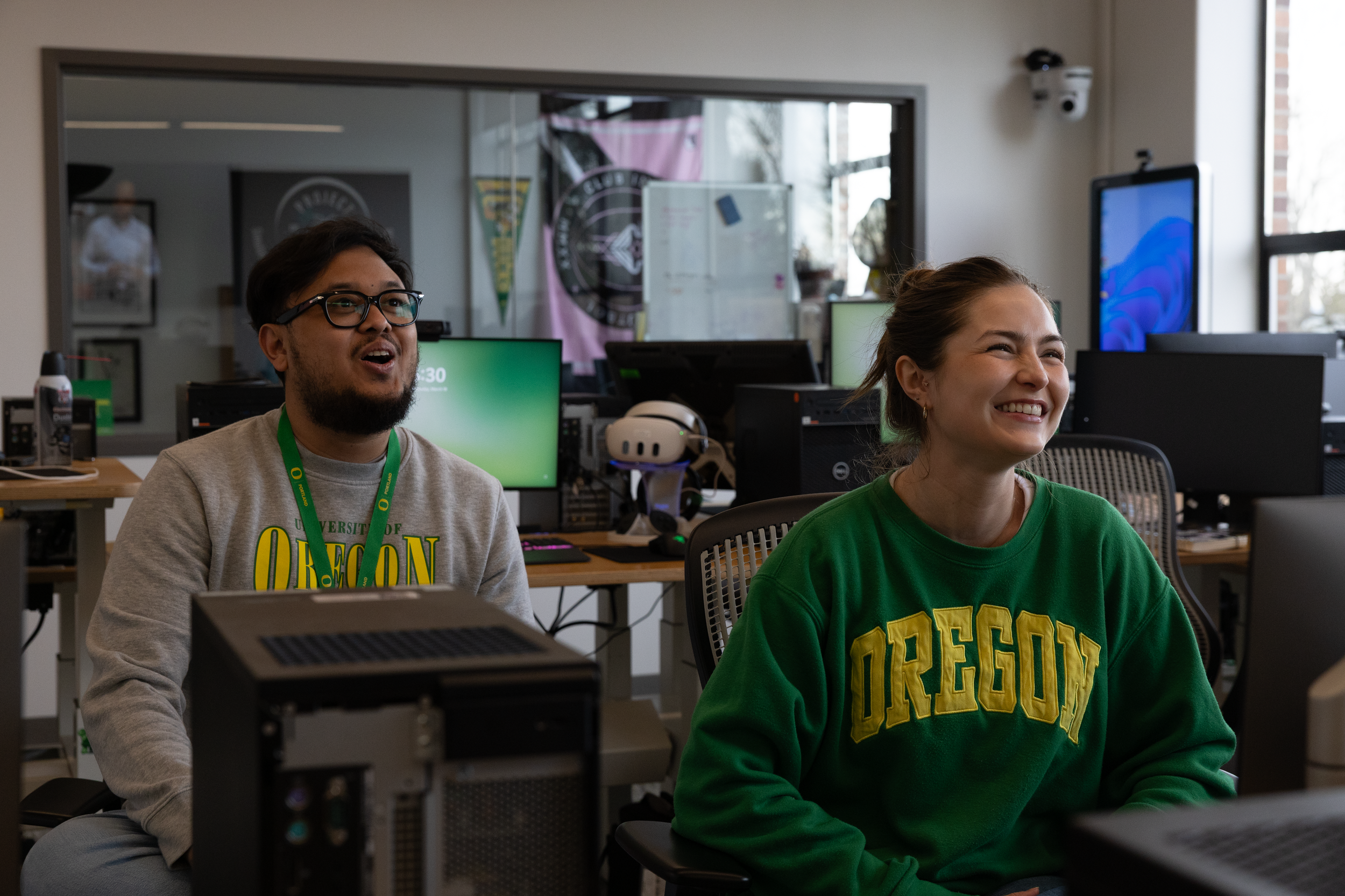 A man with glasses and a beard and a woman with her hair pulled back, smiling, sitting in an office environment at desks with computers and monitors, wearing University of Oregon sweatshirts.