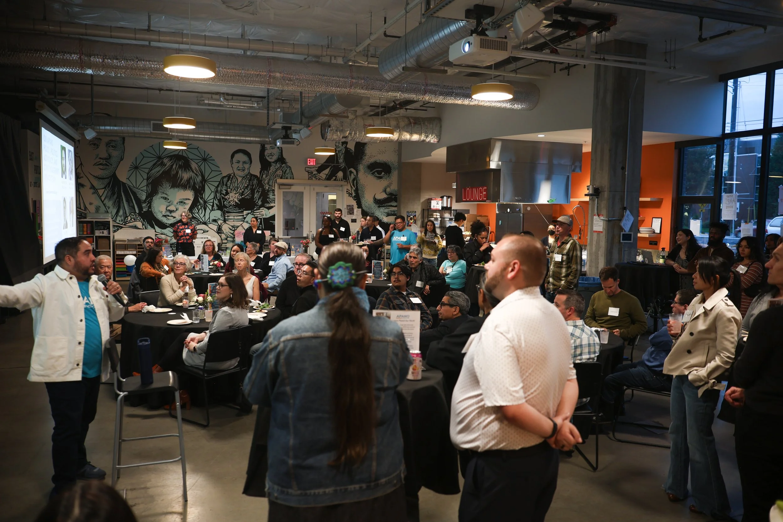 People gathered at an indoor event with a speaker in a white coat on stage, a large mural of diverse faces on the wall, and high ceilings with industrial ductwork and lighting. The room has large windows and a sign that says 'Lounge'.