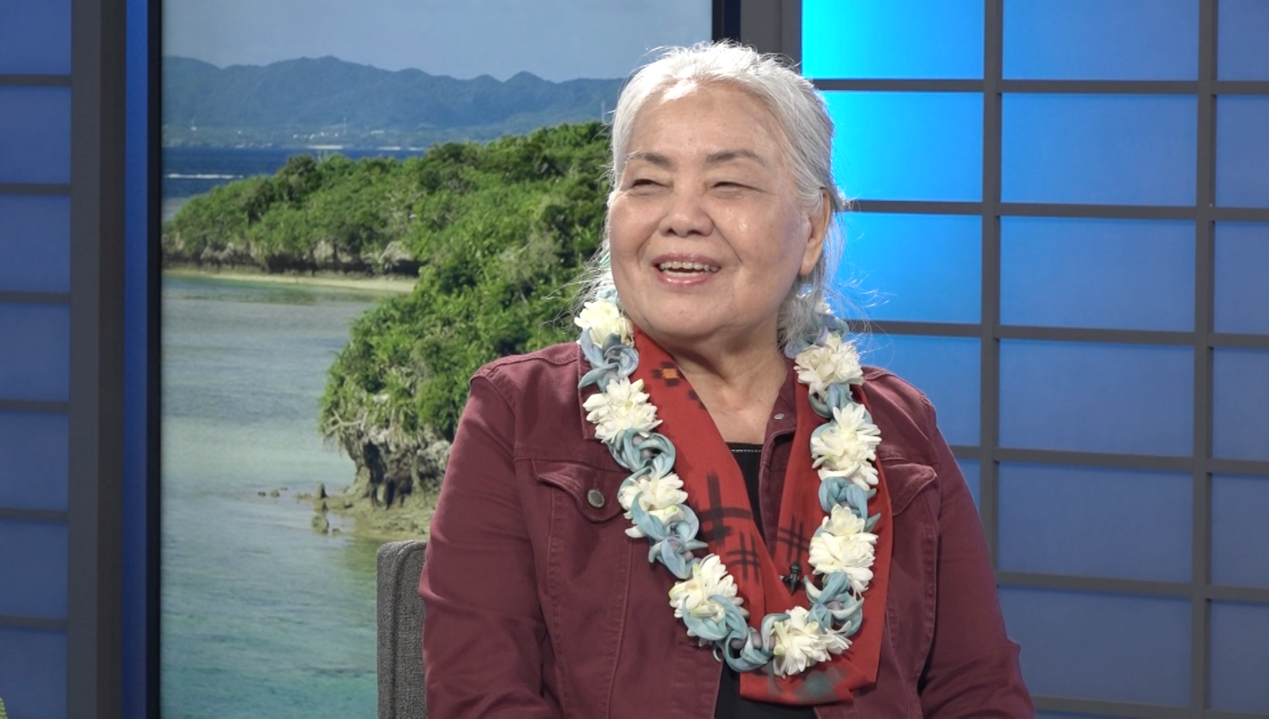An elderly woman with gray hair, wearing a reddish-brown jacket and a red and black scarf, sits in front of a scenic background of mountains, water, and greenery. She has a floral lei around her neck and is smiling.
