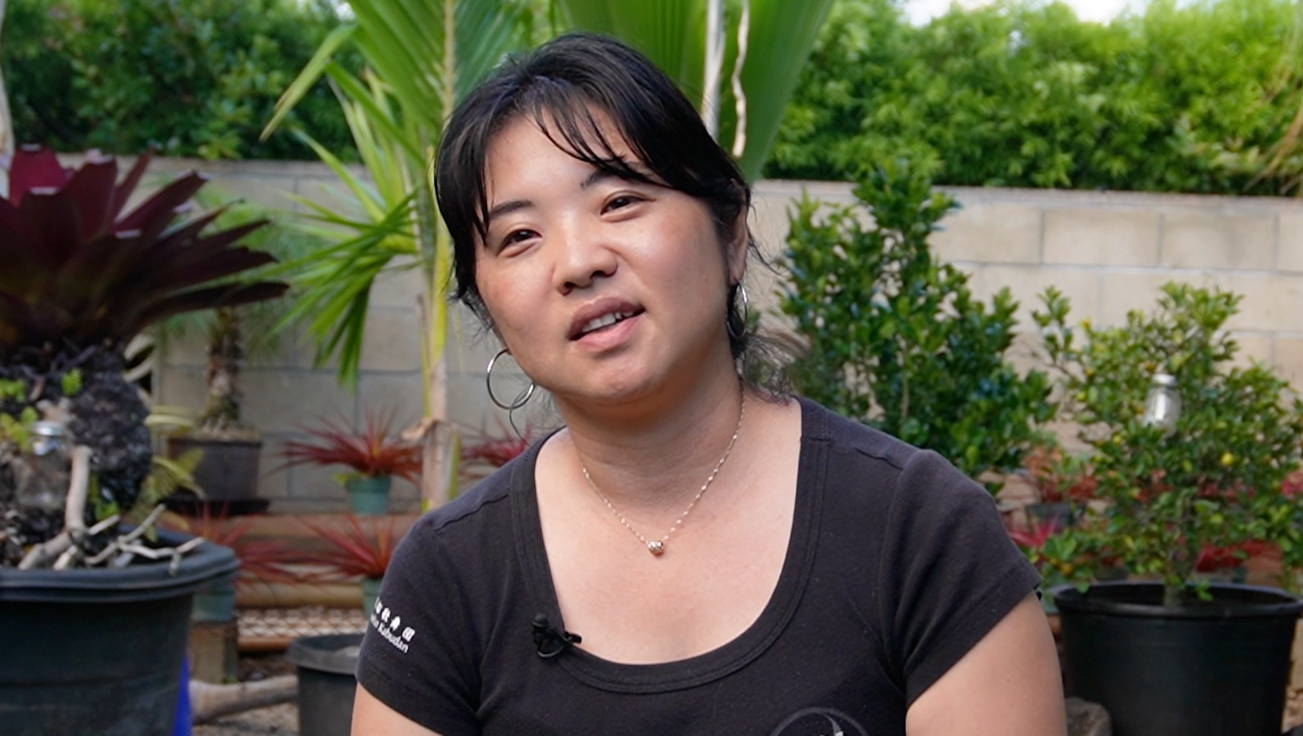 A woman with black hair wearing a black t-shirt with earrings and a necklace, sitting outdoors surrounded by potted plants and greenery.