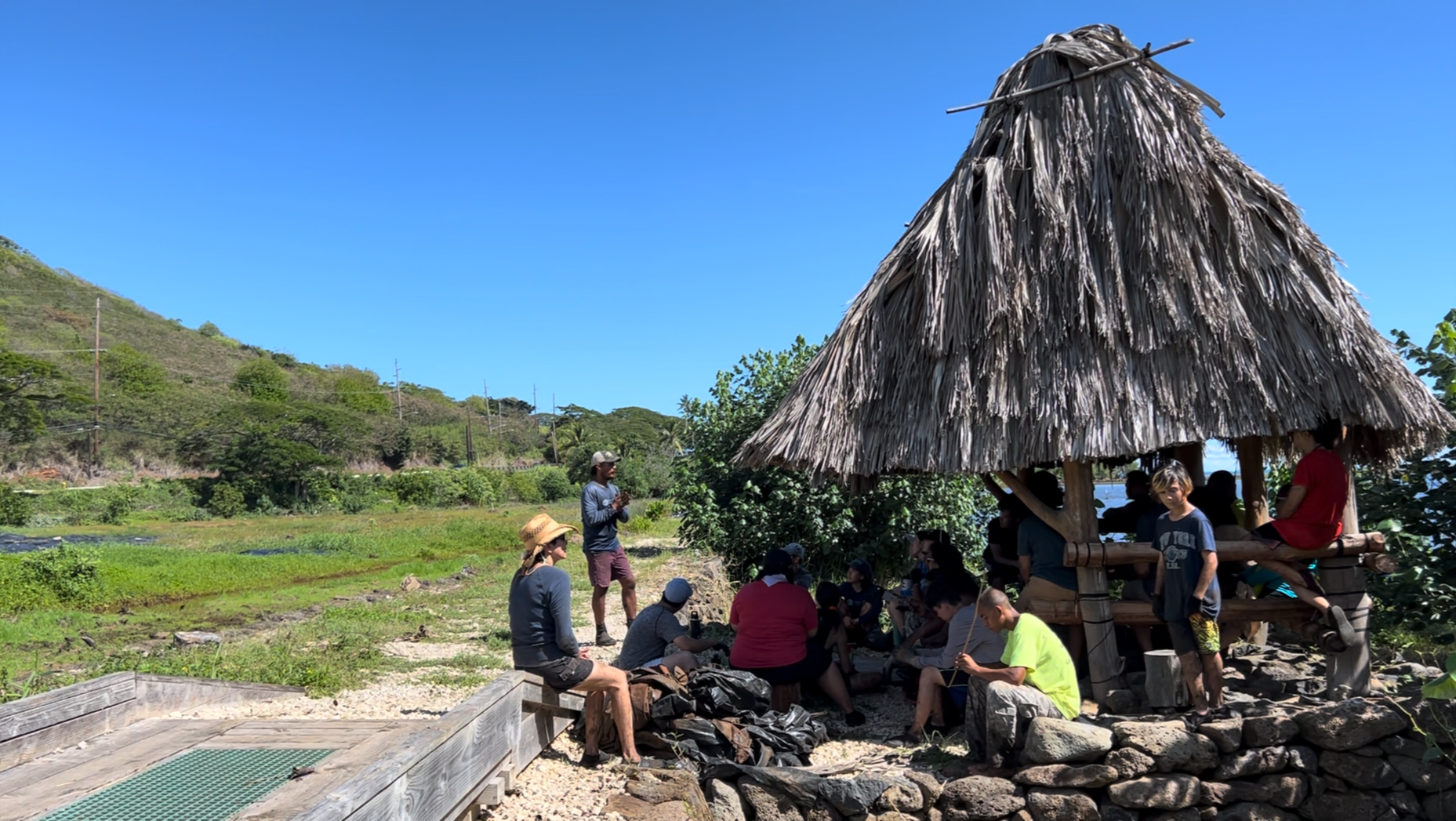 Group of people gathered under a thatched-roof structure in a lush outdoor setting with green hills and a blue sky in the background.