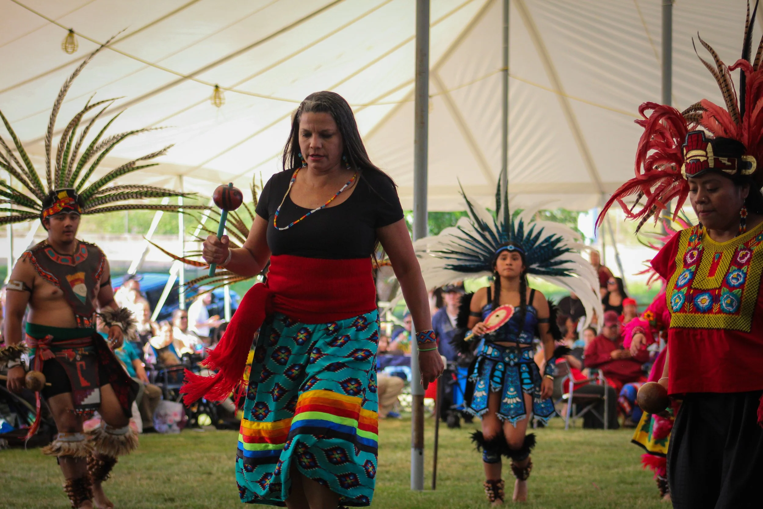 Native dancers performing at a cultural event under a large white tent, with some dressed in traditional attire with feathers and vibrant patterns, and a woman in the foreground wearing a black top and colorful skirt.