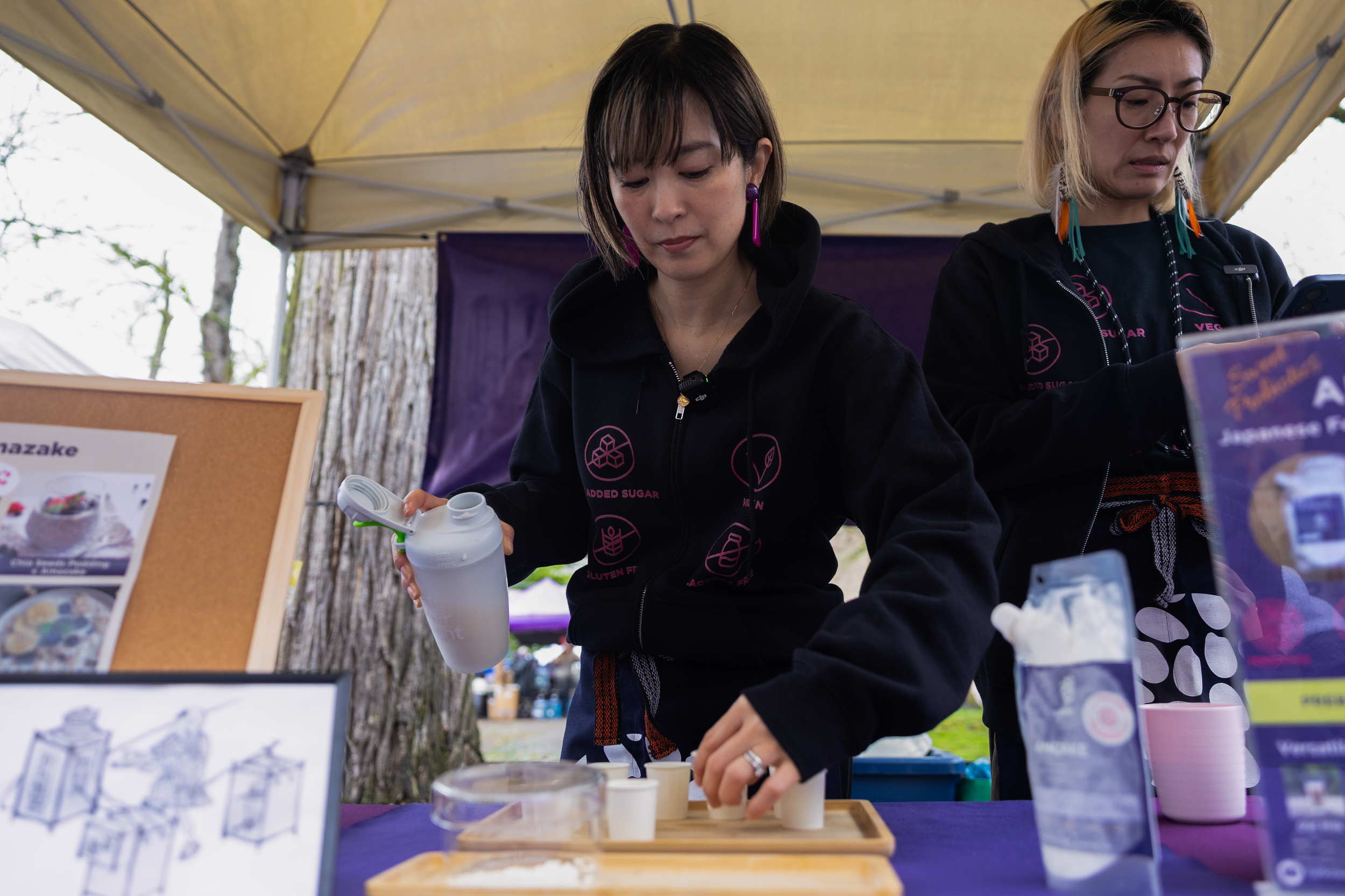 Two women are working at a booth outdoors under a yellow canopy. One woman is pouring a liquid into small cups, and the other woman is looking at her phone. There are informational signs and food packages on the table.