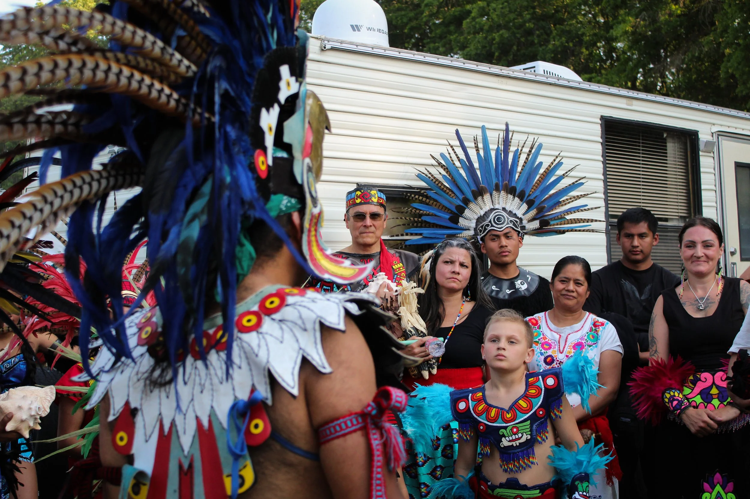 People dressed in traditional Native American costumes with feathered headdresses and colorful beadwork gather outdoors during a cultural event.