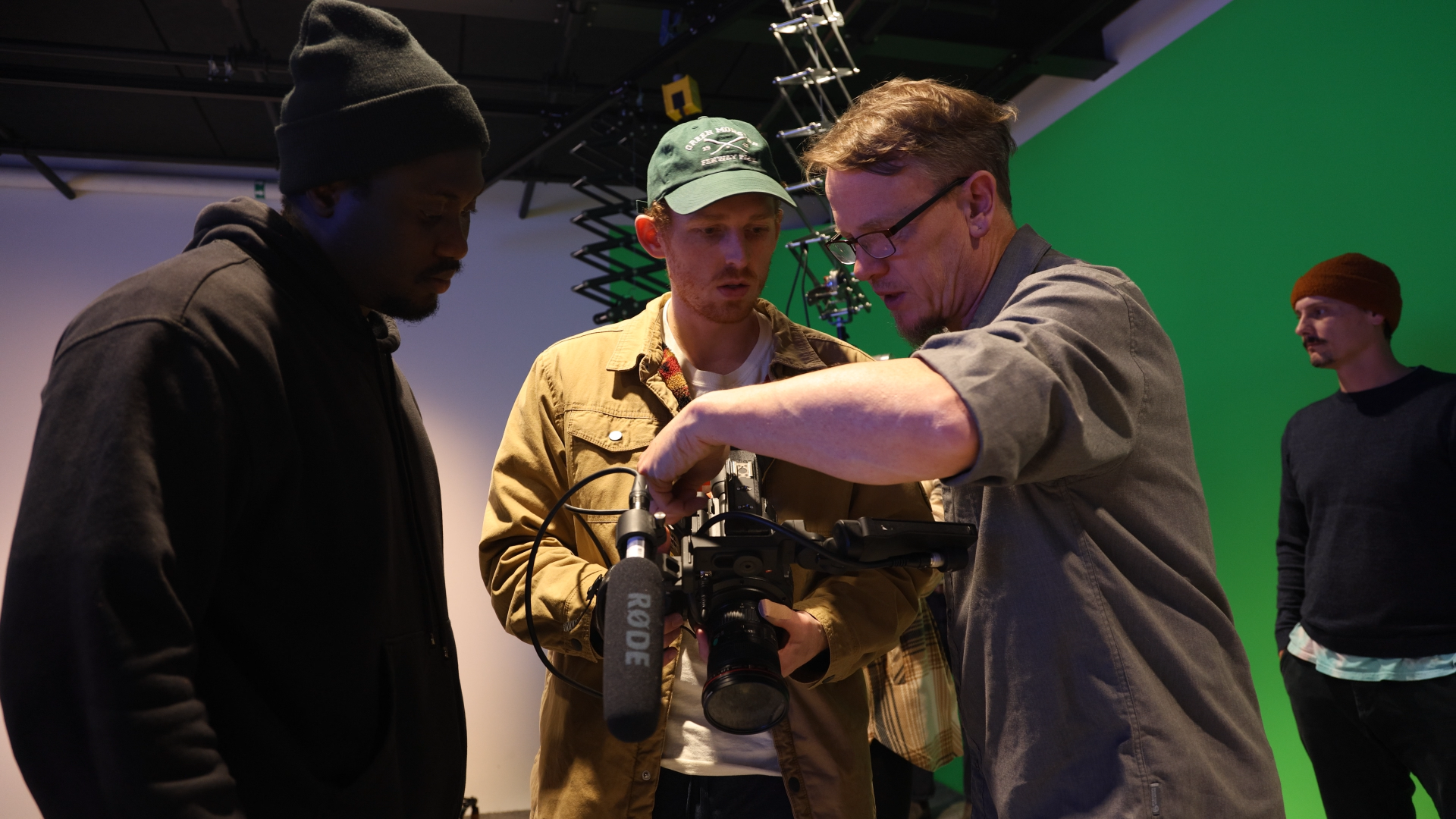Group of four men working together with a camera on a green screen background, one of them wearing glasses and a baseball cap.