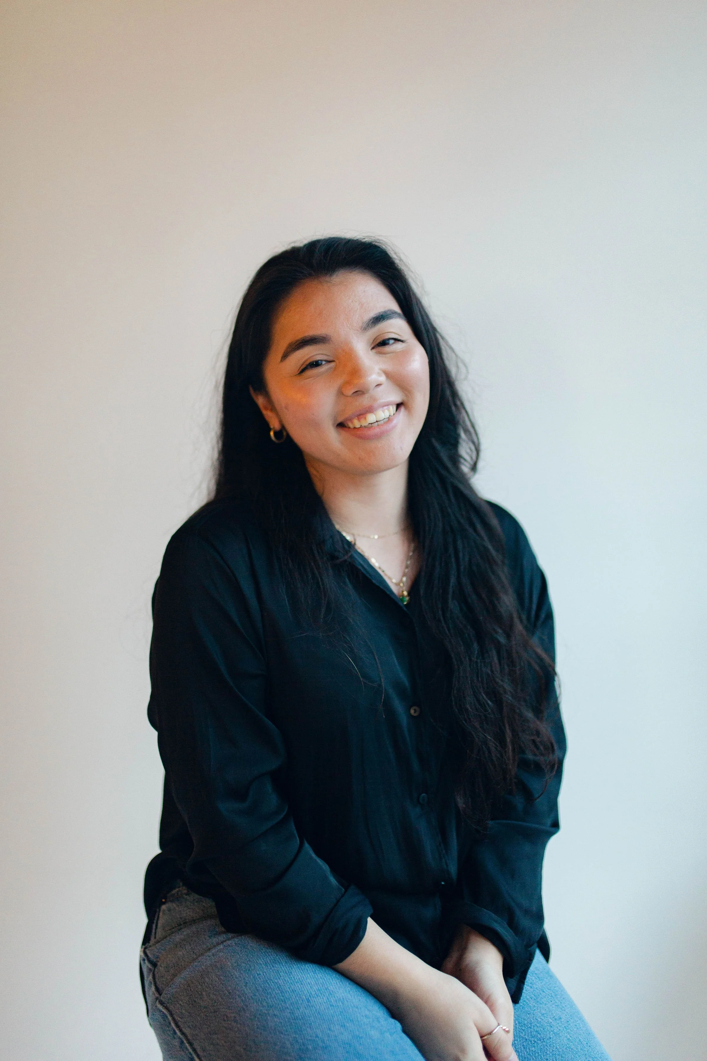 A young woman with black hair, wearing gold earrings, a black shirt, and light blue jeans, sitting against a plain light-colored wall, smiling at the camera.