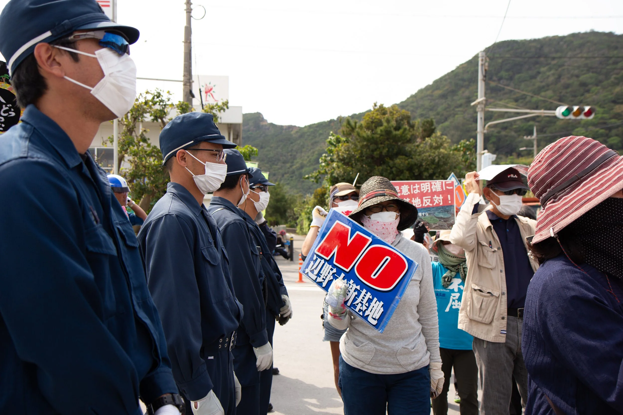 People protesting outdoors, wearing masks, holding signs with text and Japanese characters, with mountains and traffic lights in the background.