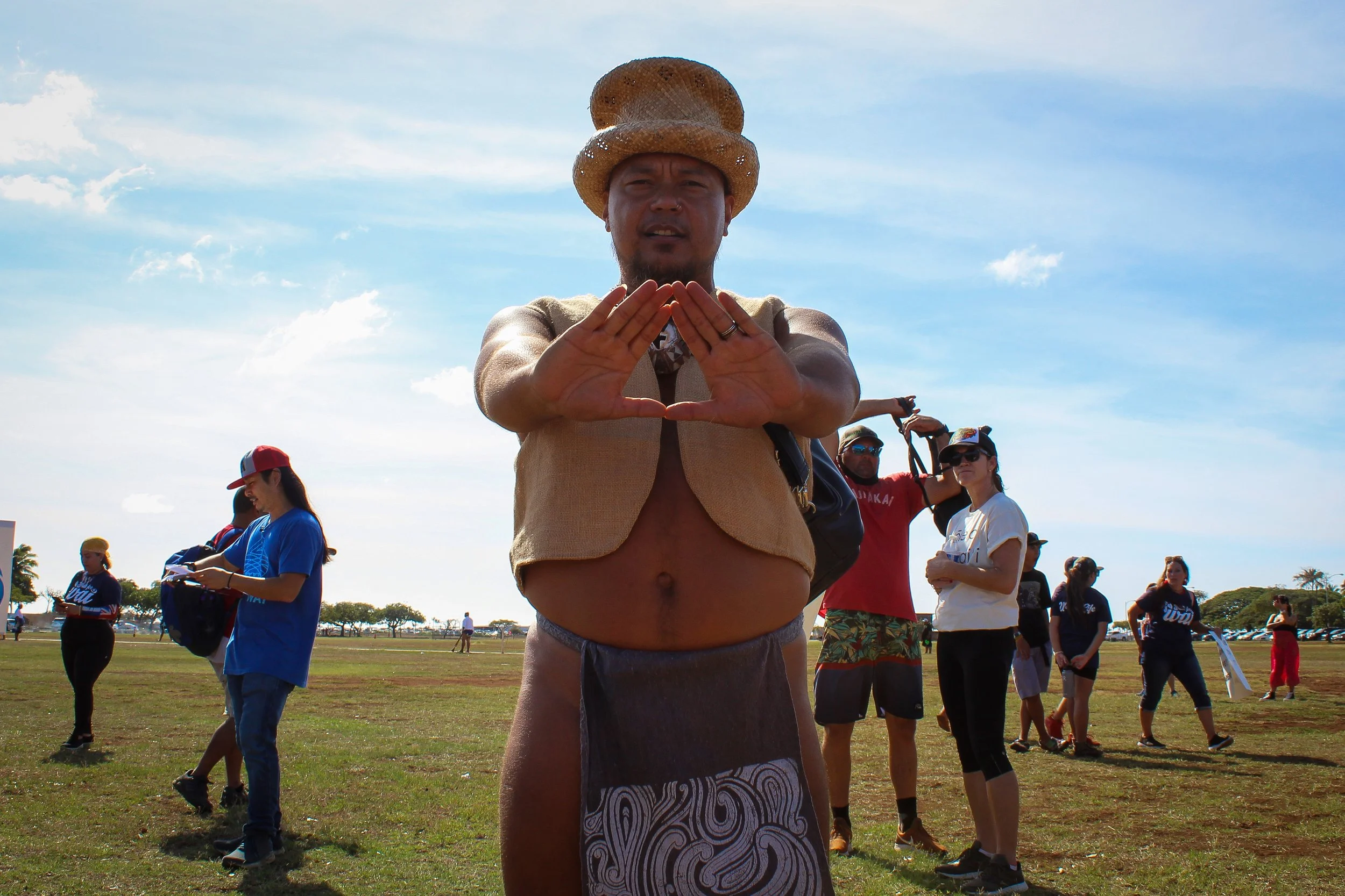 Man wearing a large straw hat making a triangle shape with his hands at a crowd gathering outdoors under a partly cloudy sky.