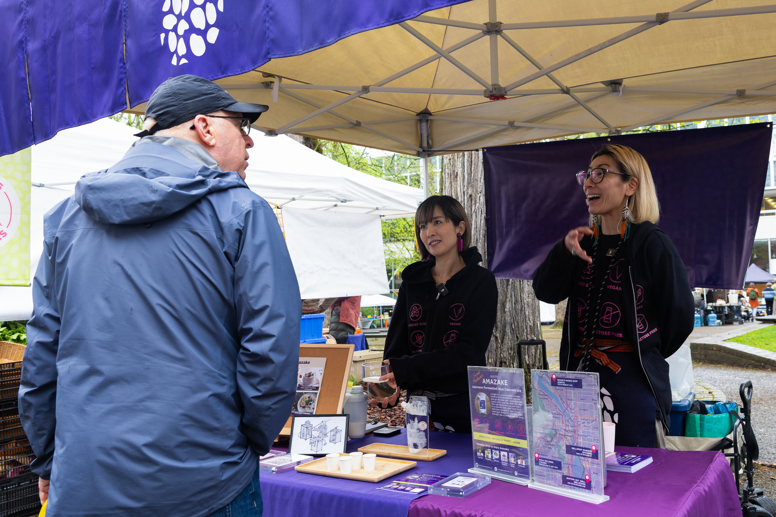Three people engaging at an outdoor event booth under tents, two women behind the booth, one man facing them. The women are wearing black hoodies with pink and white vegan and food-related icons, and the man is wearing a blue rain jacket and a black cap. The booth has informational display boards, flyers, and small samples.