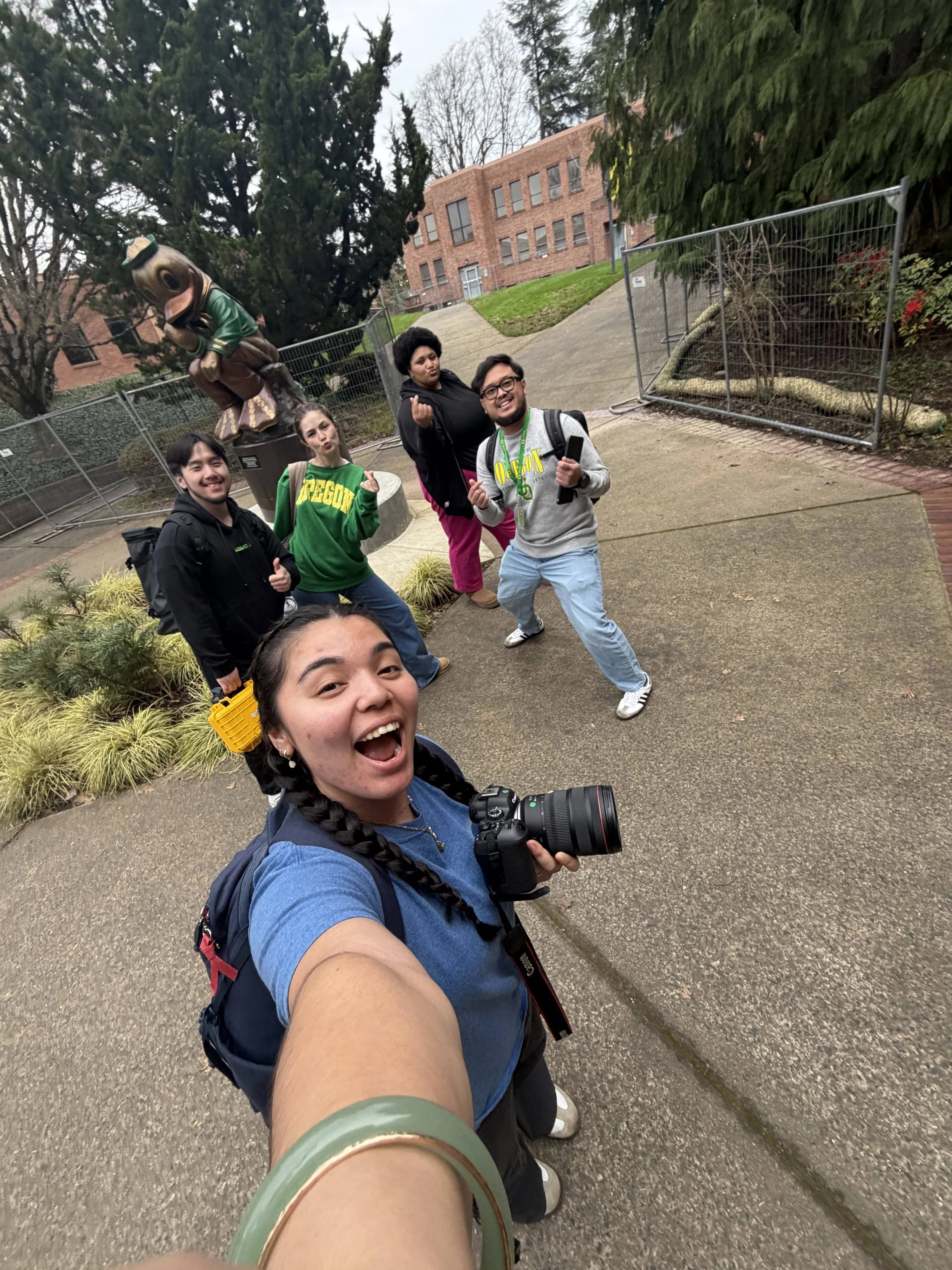 A group of five people taking a selfie outdoors. A woman in the foreground is holding a camera, and four others are smiling behind her. They are standing on a sidewalk near bushes and trees, with a brick building in the background.