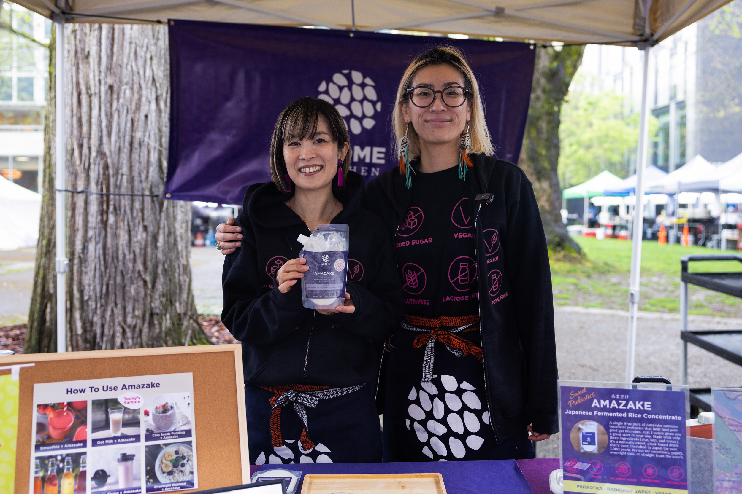 Two women standing behind a booth at an outdoor event. One is holding a package of amazake and smiling. The booth has posters with images of drinks and a purple banner in the background.
