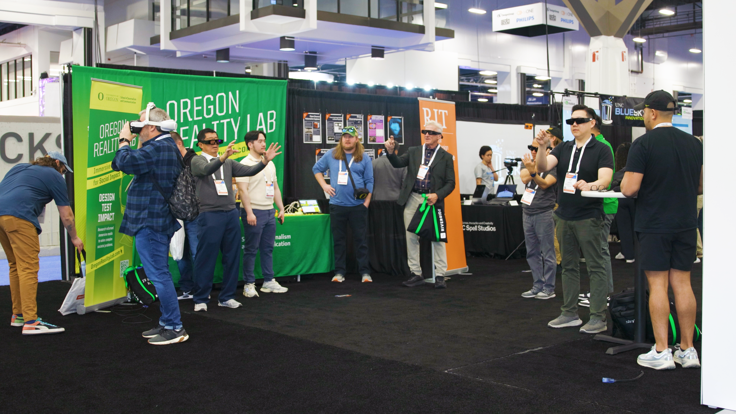 People at a technology expo standing near a green banner for Oregon Reality Lab, some wearing virtual reality headsets, engaging with each other and exploring displays in a busy indoor event space.