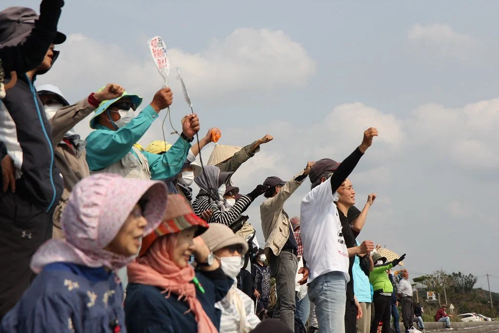 A crowd of people standing outdoors, many with raised fists, some wearing masks, hats, and jackets, participating in a protest or rally under a cloudy sky.