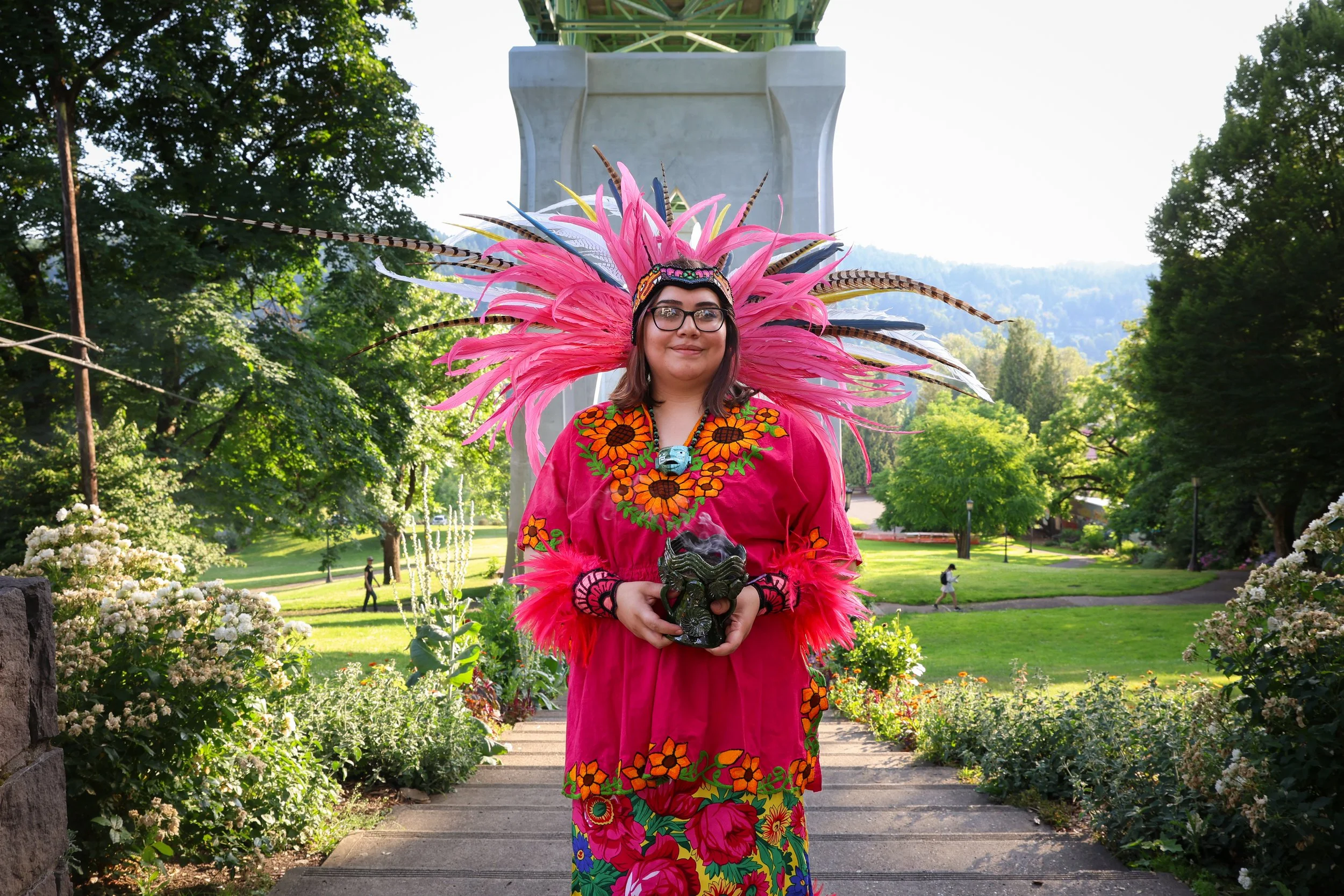 A woman wearing a colorful pink and floral traditional outfit and a large headdress with pink feathers and animal print feathers standing in a park with trees, flowers, and people walking in the background.