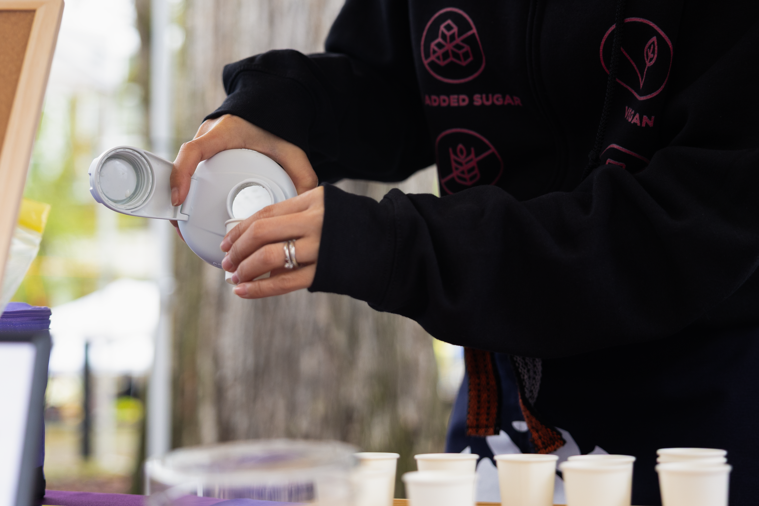 Person pouring a white liquid into small paper cups outdoors, with trees in the background.