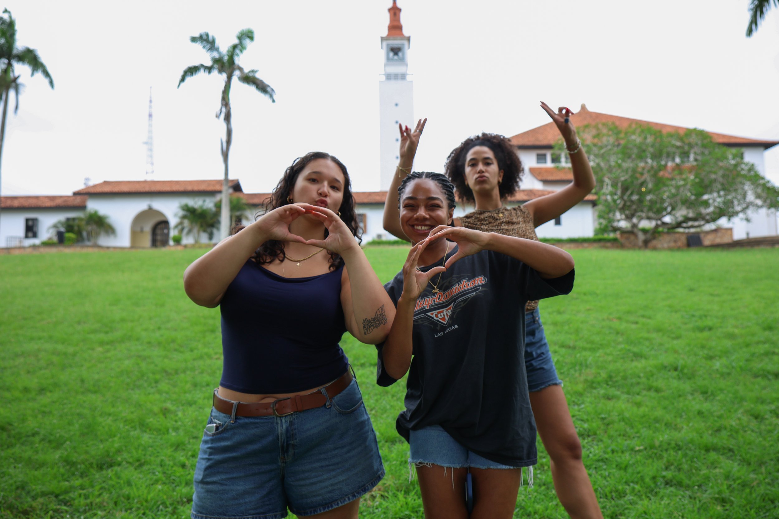 Three young women standing on a grassy lawn making heart and peace signs with their hands. Two women are smiling, while the third is making a pout. The background features a white building with a tower, palm trees, and a cloudy sky.