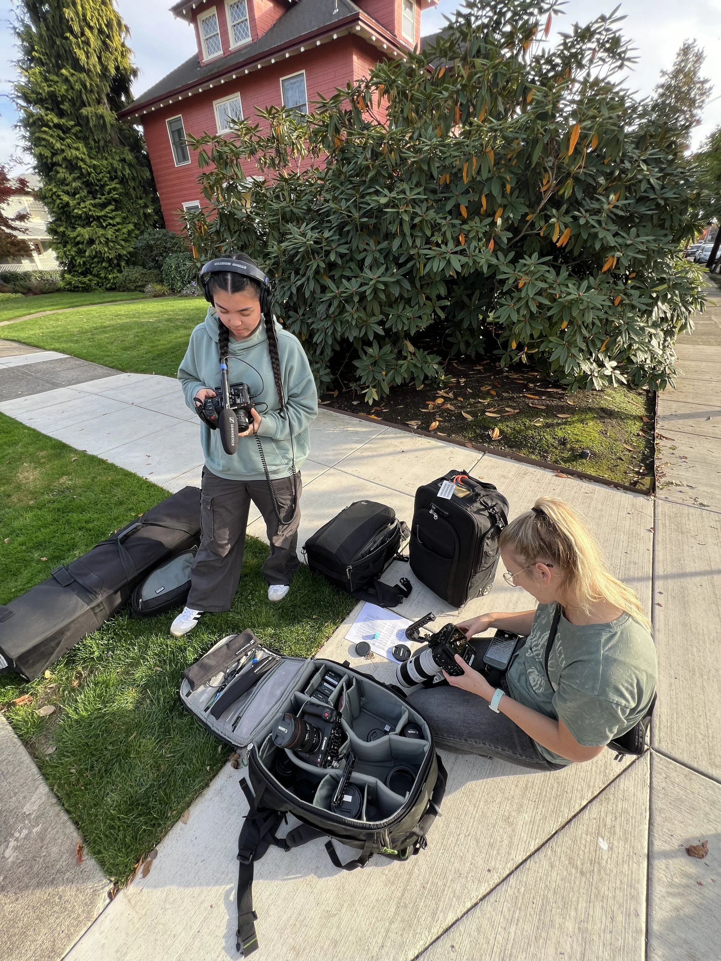 Two females preparing camera equipment outdoors on a sidewalk near a grassy area and a large shrub. One woman with dark braided hair and a gray hoodie is standing and looking at a camera, while the other woman with blonde hair and glasses is sitting and adjusting a camera in her hands. Camera bags and gear are laid out on the ground.