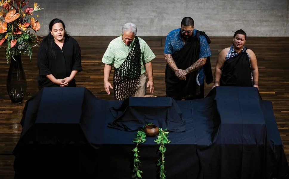 Four people stand solemnly behind a table draped with black cloth, with a small pot and greenery as memorial objects, in a dimly lit room with wood flooring and a floral arrangement to the side.