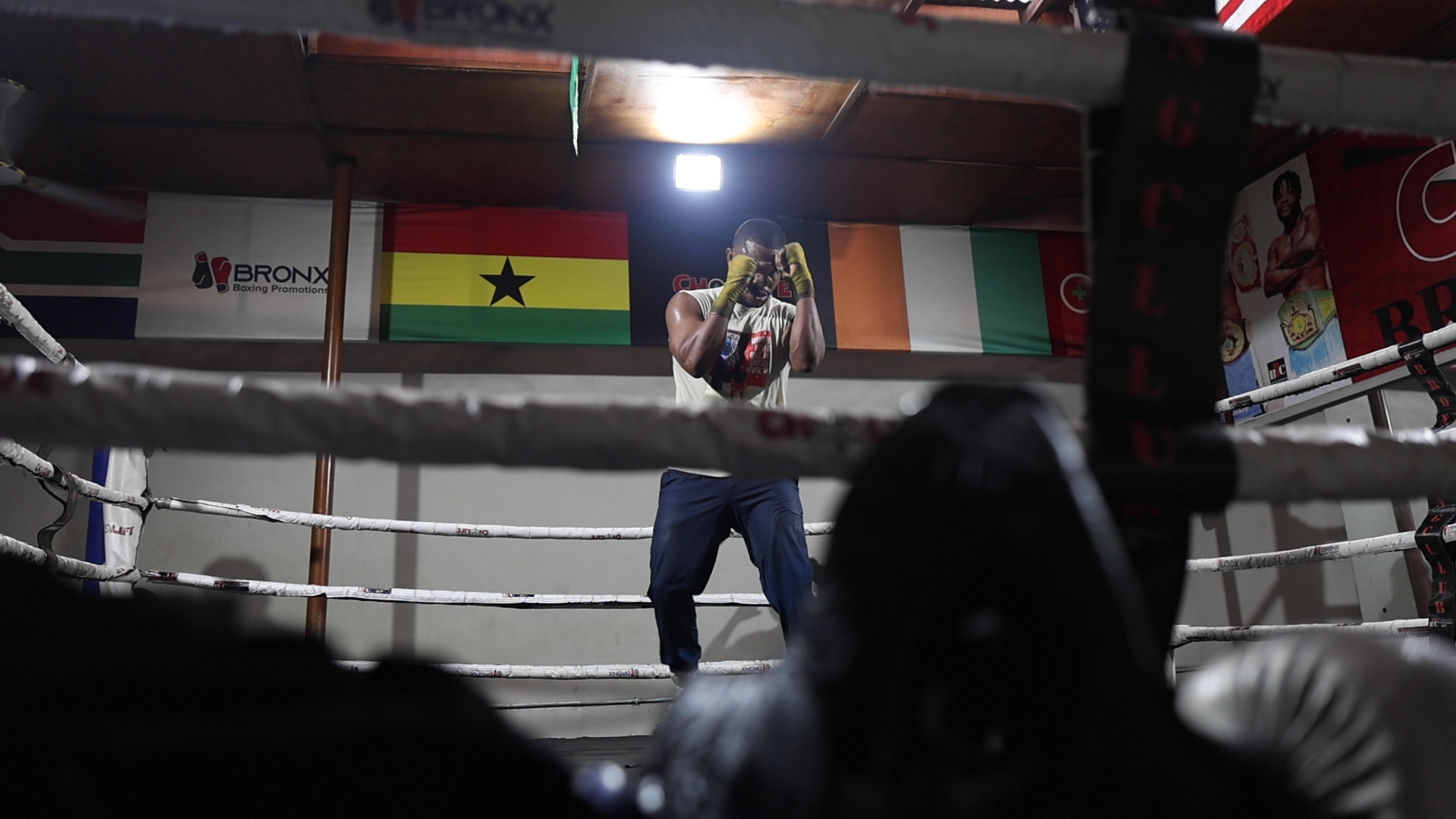 A boxer in a gym, standing in the ring with his hands on his head, viewed from outside the ring through the ropes. Flags of Ghana, Ireland, and another country are hanging behind him.