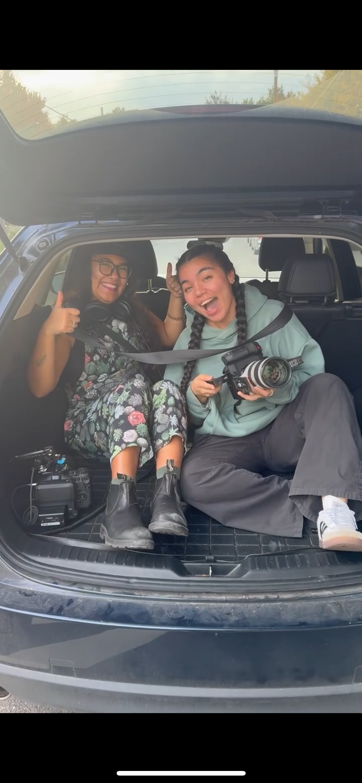 Two young women sitting in the open trunk of a car, smiling and giving thumbs-up. One woman has braided hair and is holding a professional camera; the other woman wears glasses and headphones around her neck. Both appear to be happy and enjoying a moment together.