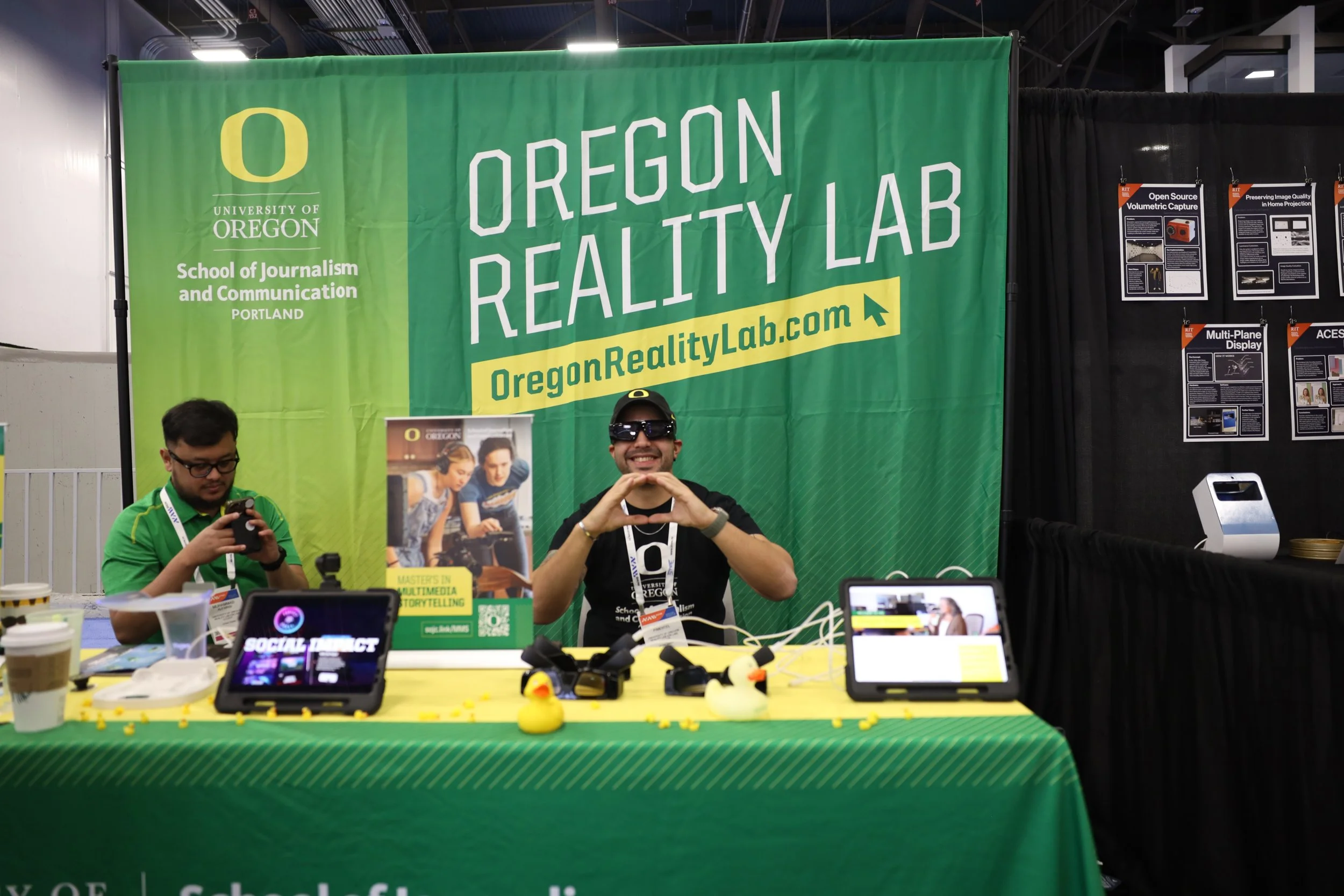 A booth at an event for the University of Oregon's School of Journalism and Communication, featuring a green banner that reads "Oregon Reality Lab" and a website address. Two men are at the booth: one is using a smartphone, and the other is smiling, 