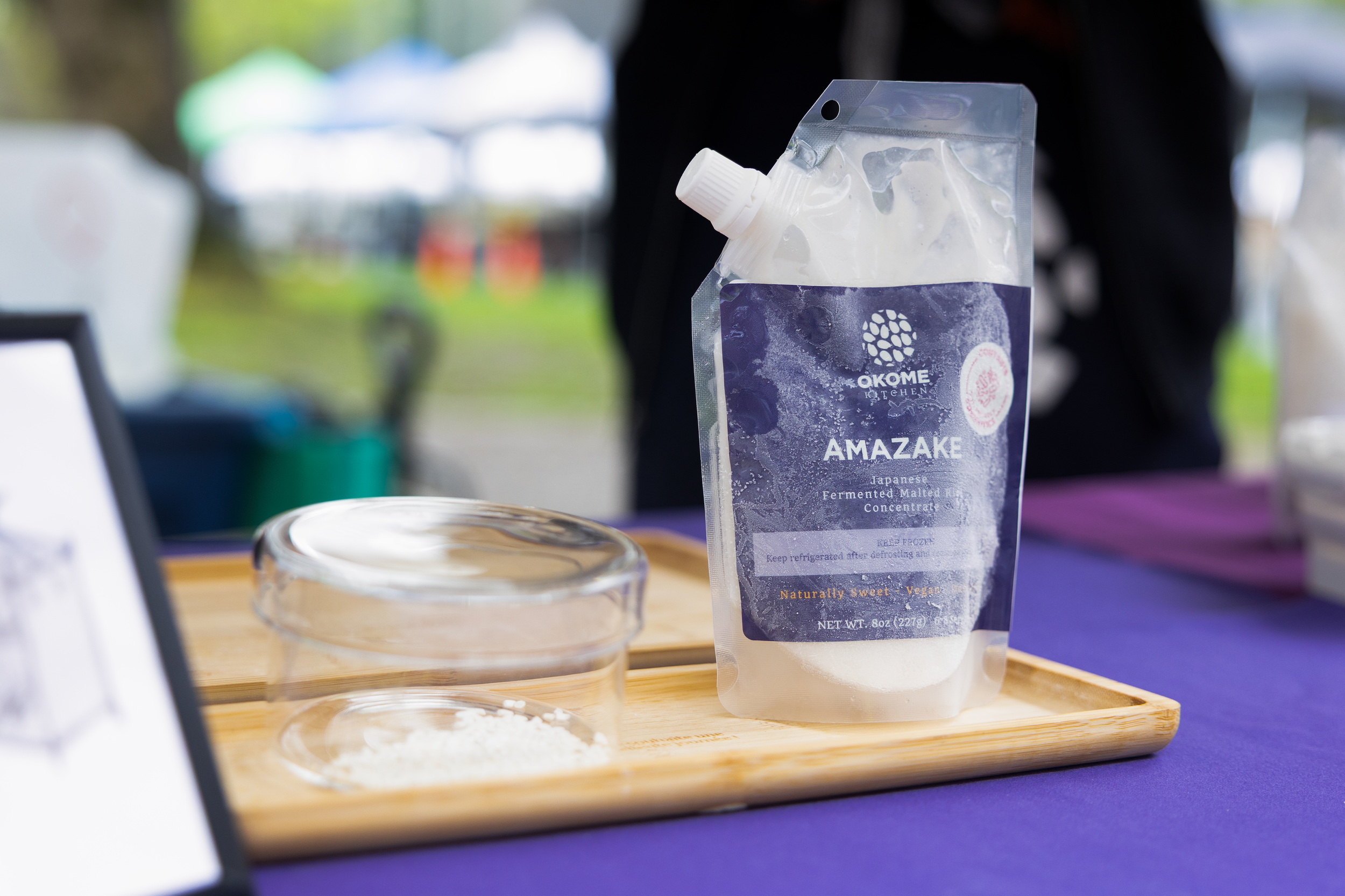 A stand-up pouch labeled 'Amazake' from Okomé Kitchen, containing Japanese fermented malt rice concentrate, placed on a wooden tray with small bowls of powder or powder-like ingredients nearby. The background is blurry with some outdoor elements and people.