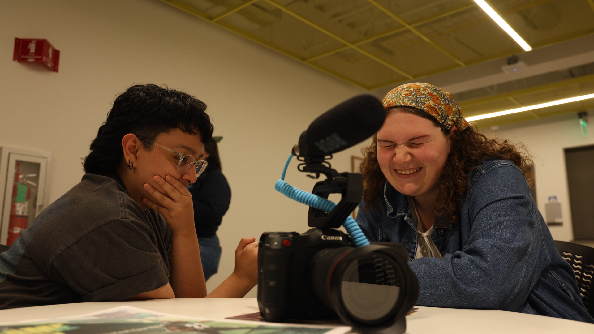 Two women sitting at a table, looking at a camera with a microscope attached. One woman, with dark hair, glasses, and earrings, appears surprised or amused, covering her mouth with her hand. The other woman, with curly hair and a colorful headscarf, 