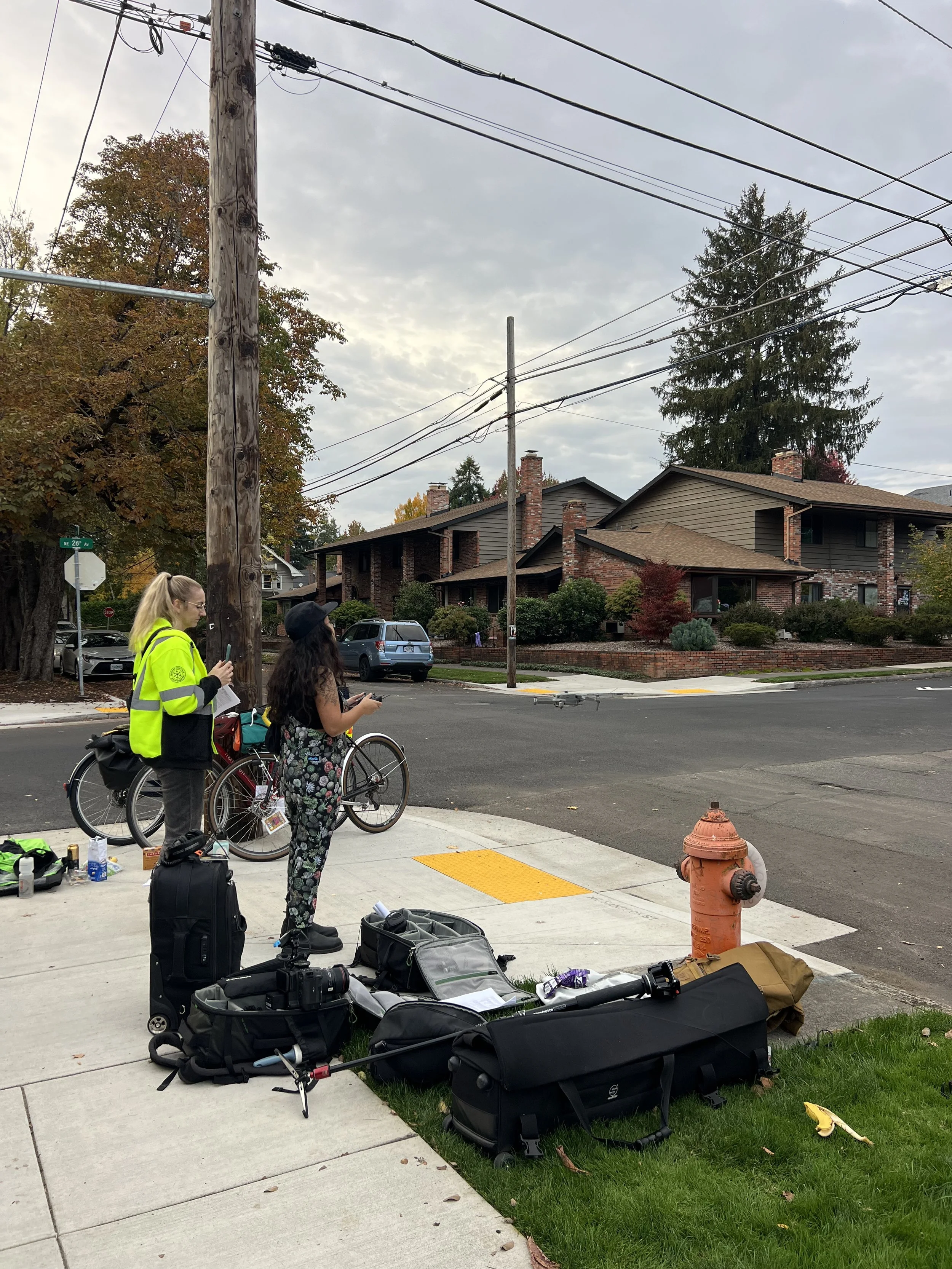 Two women standing on a sidewalk with bikes and equipment laid out on the ground in front of them. One woman is wearing a yellow jacket, and the other has long dark hair. The scene is in a residential neighborhood with trees and houses in the background, overcast sky.