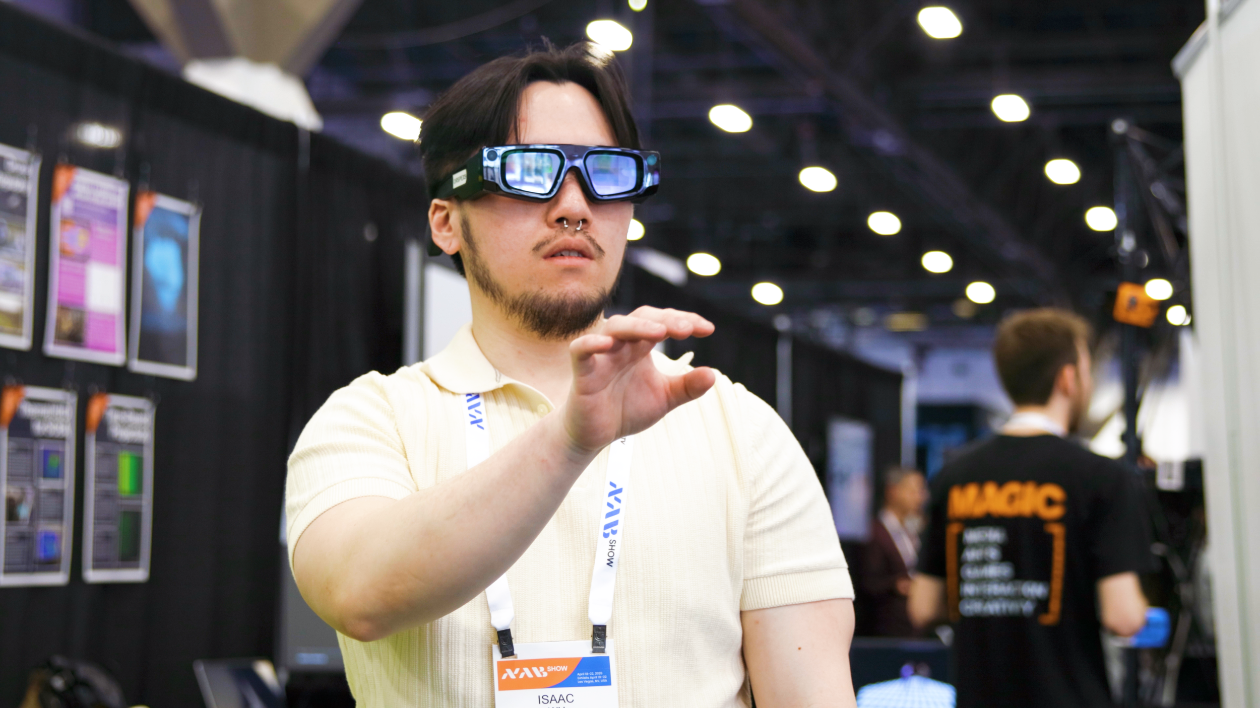 A man wearing Smart glasses at a technology convention or trade show, with a black lanyard and a white badge that says 'Isaac' and 'MIX Show' visible around his neck, standing in front of display panels and screens.