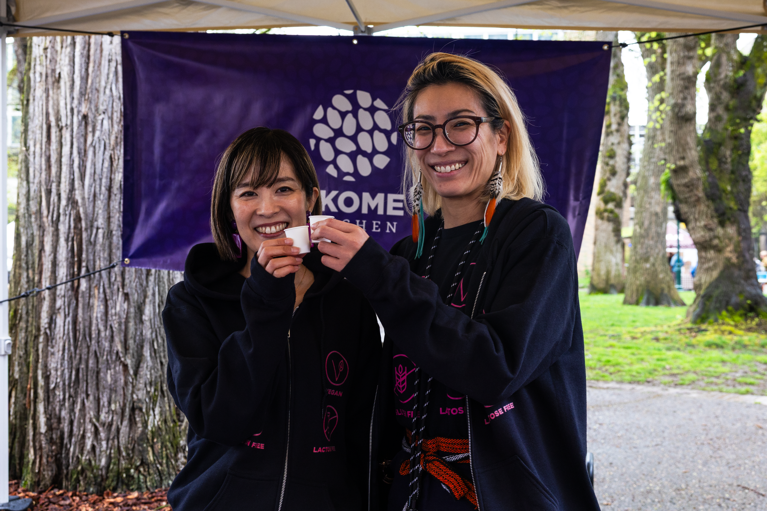 Two women smiling and clinking small cups together outdoors, with a purple banner behind them and trees in the background.