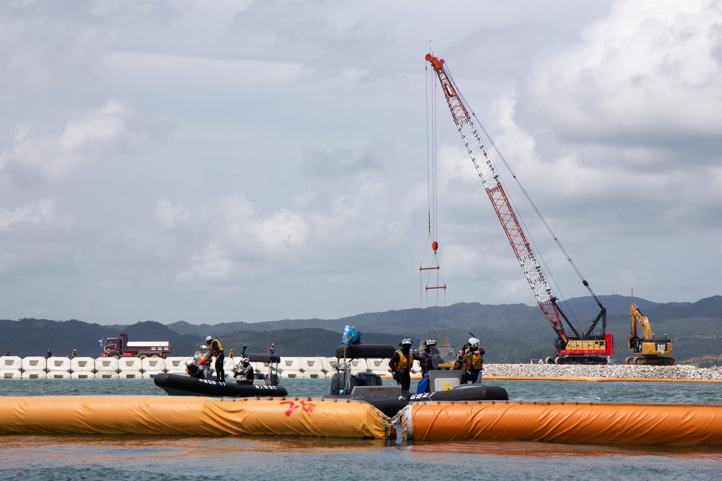 Workers in safety gear and boats on water at a construction site with a tall red and white crane against cloudy sky and distant mountains.