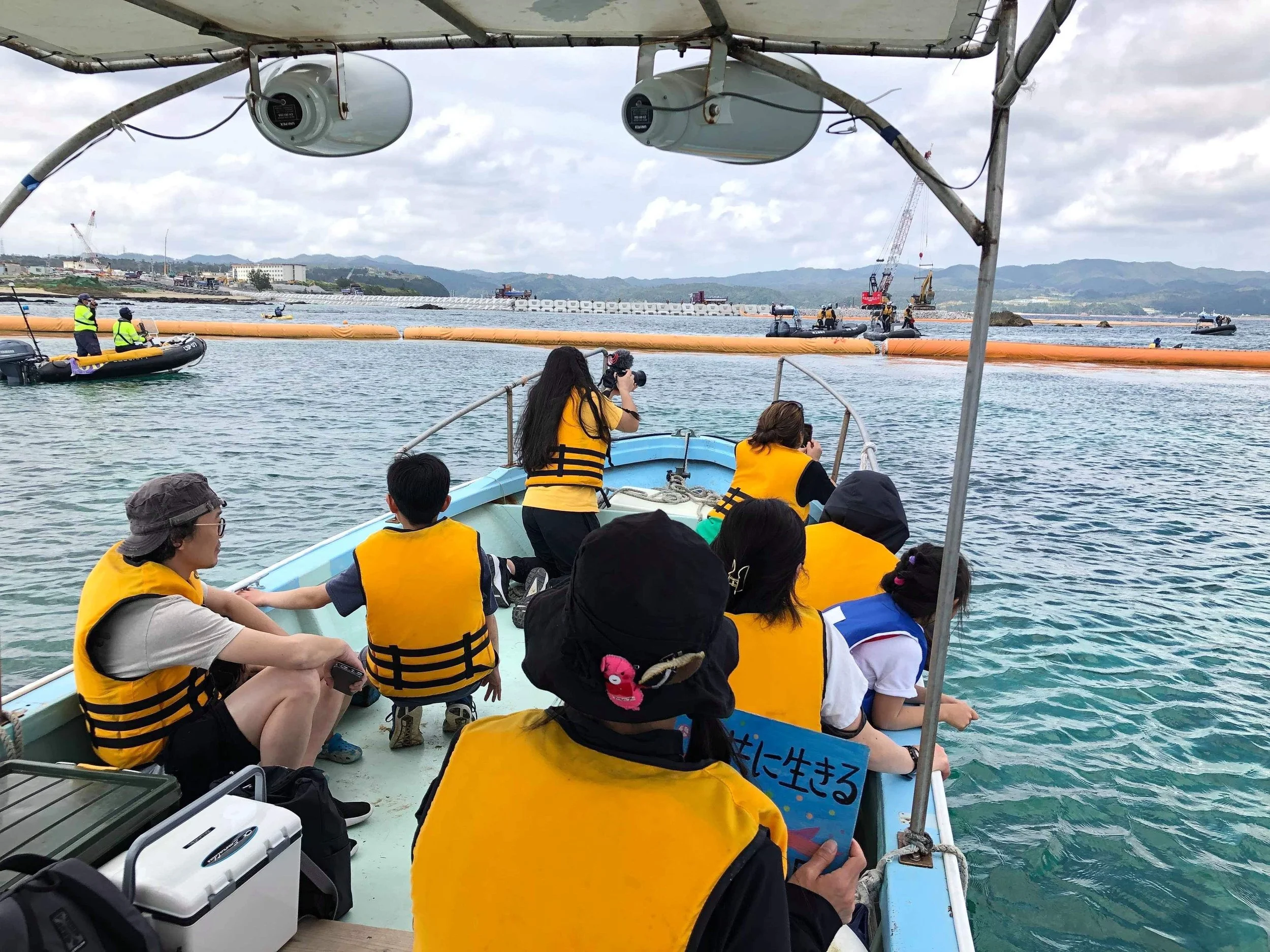 People on a boat wearing yellow life jackets, near orange floating barriers and a shoreline as they watch Japanese riot police from the ocean.