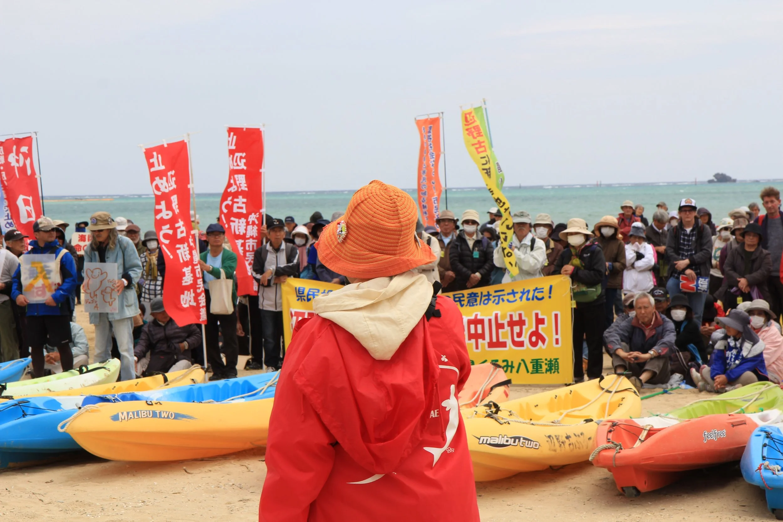 A person wearing an orange hat and red jacket facing a crowd at the beach with colorful kayaks and banners in Japanese.