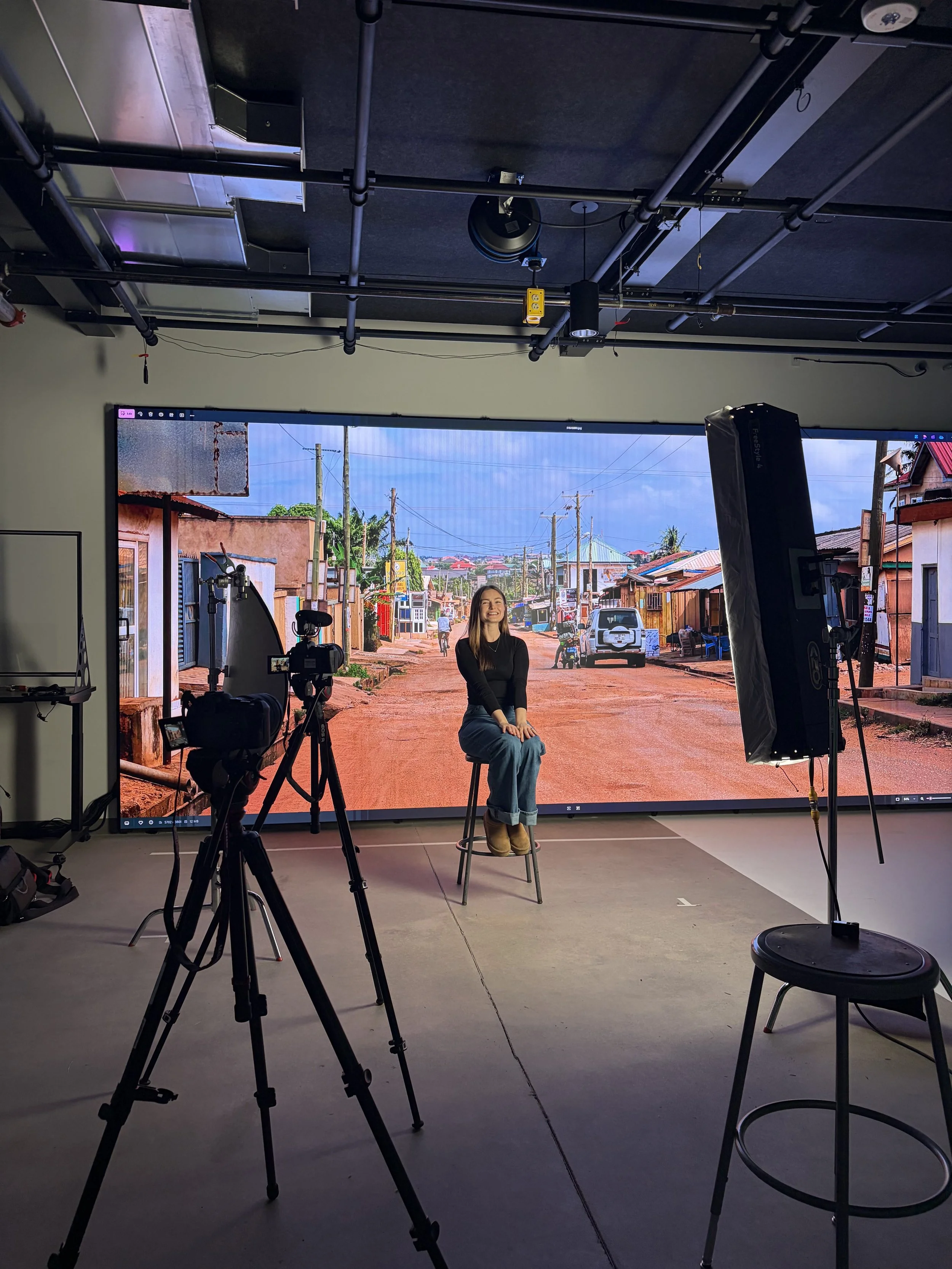 A woman sitting on a stool in a photography studio, with a large screen behind her displaying a street scene with houses, cars, power lines, and a blue sky. Studio equipment, including cameras and lights, are set up around her.