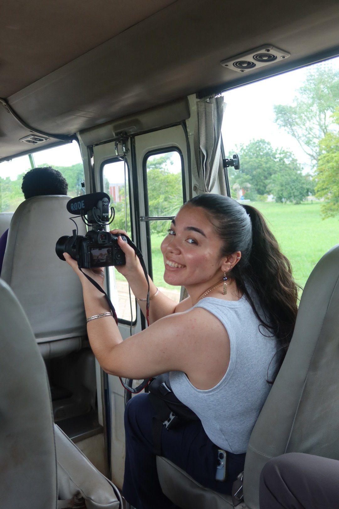 Kaiya sits in a vehicle holding a camera and smiling at the camera. She has long dark hair tied back, wearing a sleeveless gray top, earrings, and bracelets.