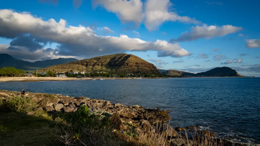 A scenic coastal view with a rocky shoreline, calm blue water, a small town or village along the beach, and a mountain in the background under a partly cloudy sky.