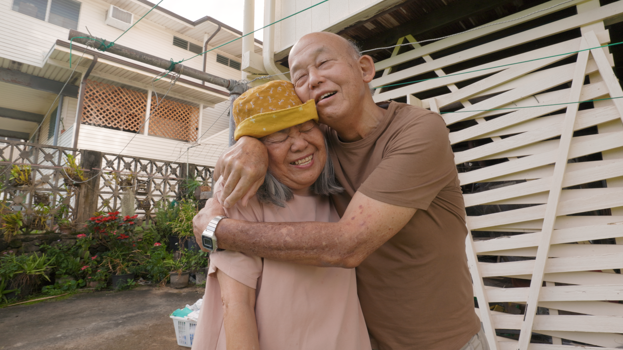 A touching moment of affection between an elderly woman wearing a yellow hat and her husband embracing her outdoors, with a house, garden, and fence in the background.
