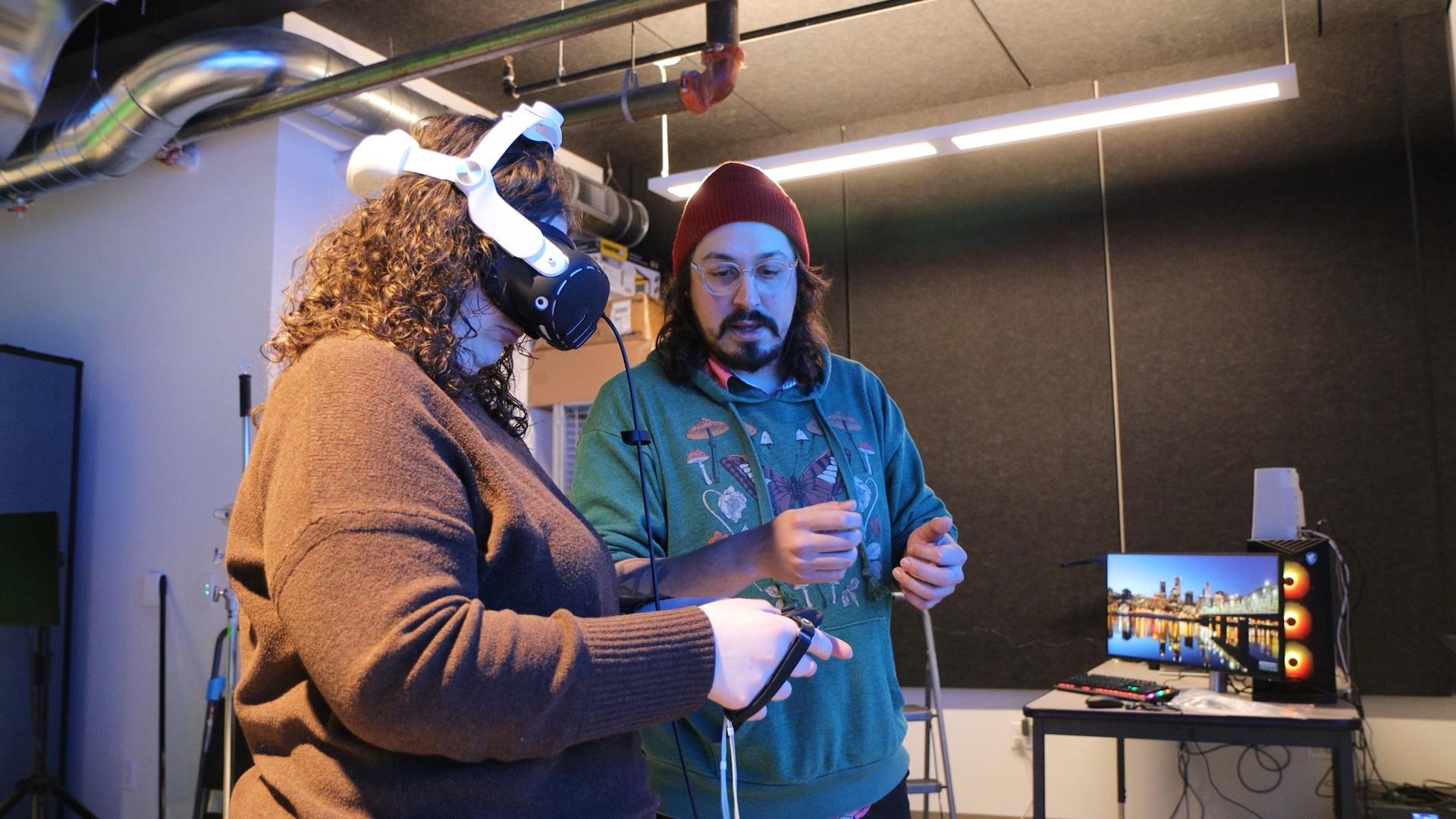 Two people in a tech lab, one with a VR headset and the other explaining, with monitors and equipment in the background.