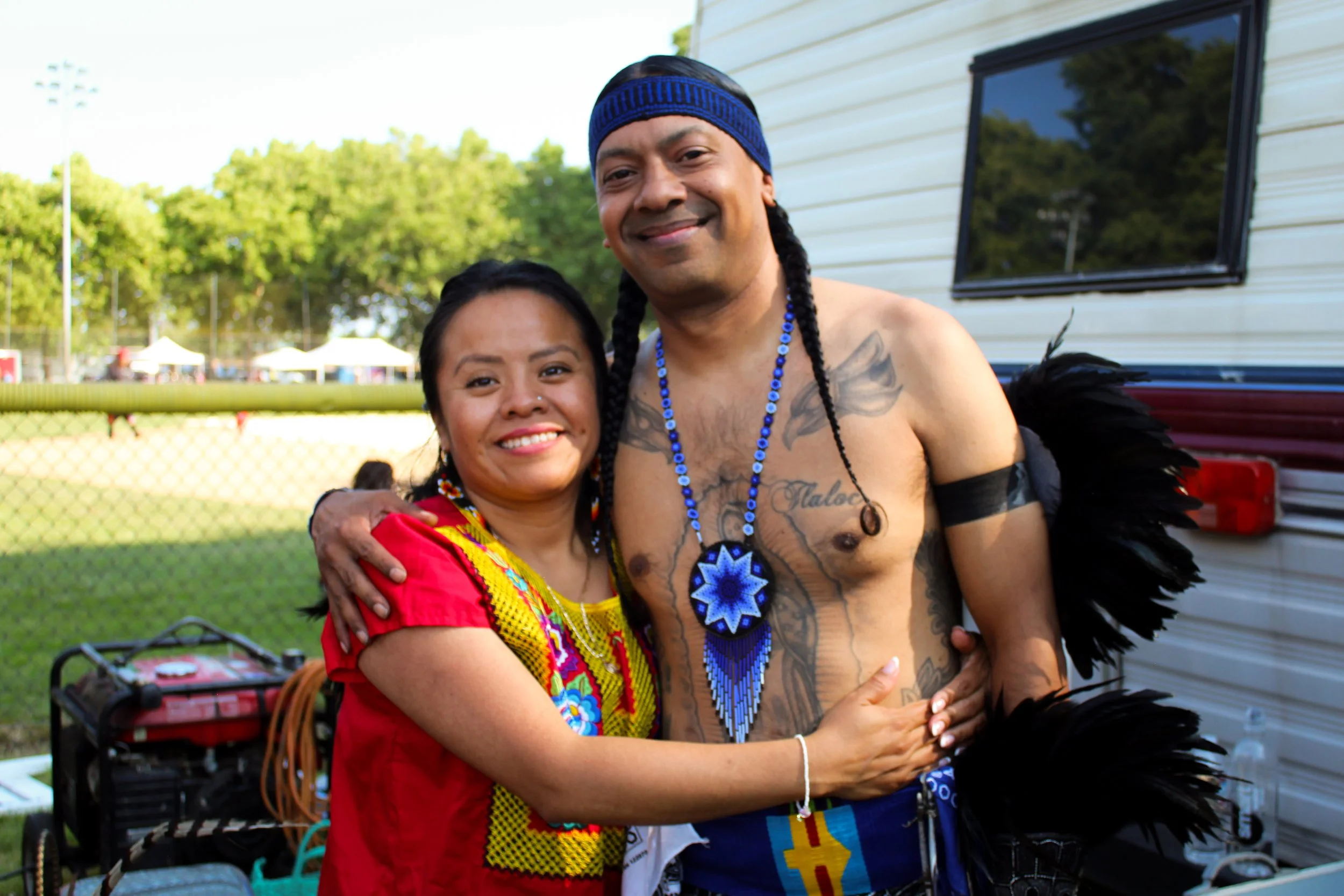 A woman and a shirtless man with tattoos and feathers, both smiling and embracing outdoors during daytime.